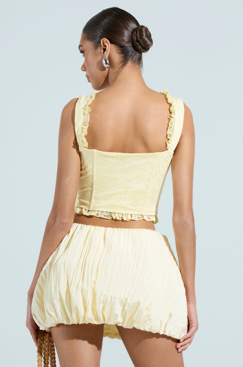 A woman with braided hair showcases a yellow SWEETHEART LACE CORSET top paired with a matching short skirt. Facing away to reveal the open back, she accessorizes with hoop earrings and carries a bag featuring a satin-lined interior.