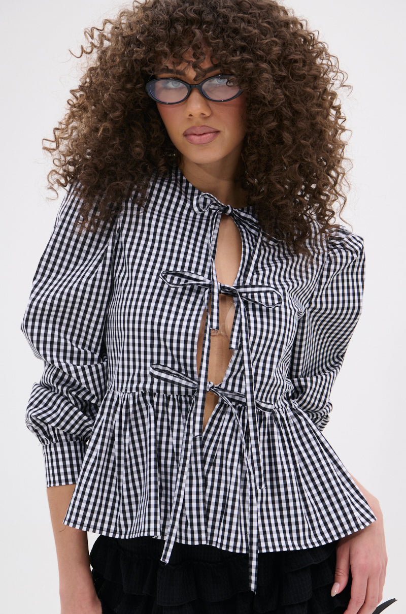 A woman with curly hair and glasses wears the LOTS OF LOVE TO GIVE GINGHAM TIE FRONT BLOUSE with puffed sleeves, bow ties, front cutouts, and a pleated peplum hem, styled with a black skirt against a plain light background.