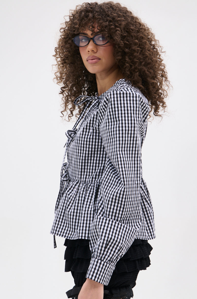 A person with curly hair and cat-eye glasses wears the LOTS OF LOVE TO GIVE GINGHAM TIE FRONT BLOUSE with bows, paired with layered black ruffle shorts featuring a pleated peplum hem, against a plain light background in lightweight cotton-blend.