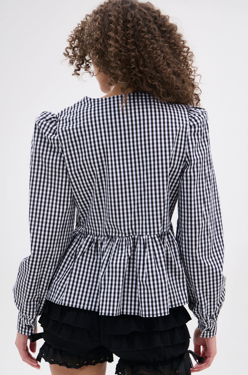 A person with curly hair, facing away from the camera, wears the LOTS OF LOVE TO GIVE GINGHAM TIE FRONT BLOUSE with a pleated peplum hem and puffed shoulders, paired with black ruffled shorts.