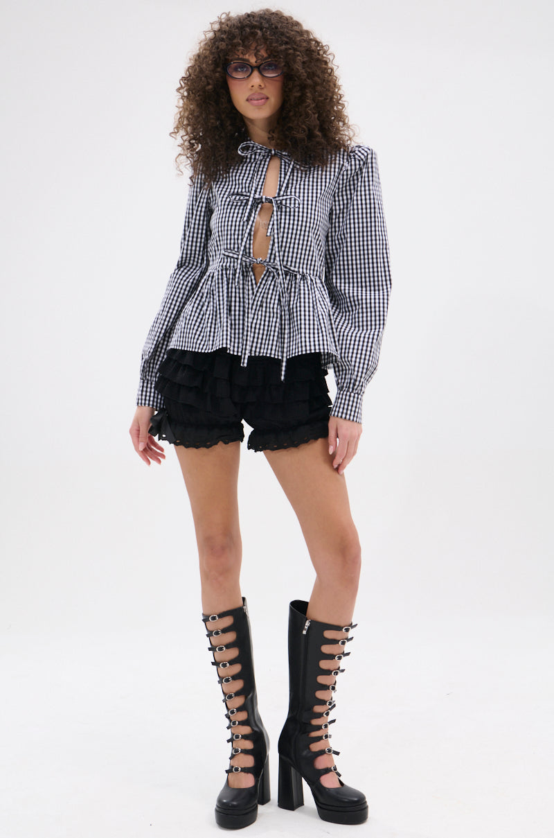 A person with curly hair and glasses wears the LOTS OF LOVE TO GIVE GINGHAM TIE FRONT BLOUSE with cutouts, black ruffled shorts with a pleated peplum hem, and tall black platform boots, standing against a plain white background.