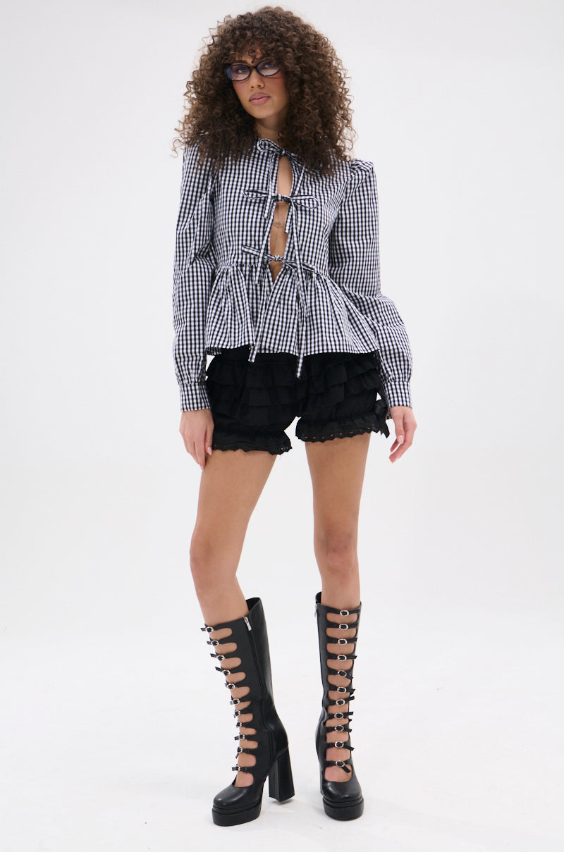A person with curly hair and glasses wears the LOTS OF LOVE TO GIVE GINGHAM TIE FRONT BLOUSE, black ruffled shorts, and tall black lace-up platform boots while standing against a plain white background.