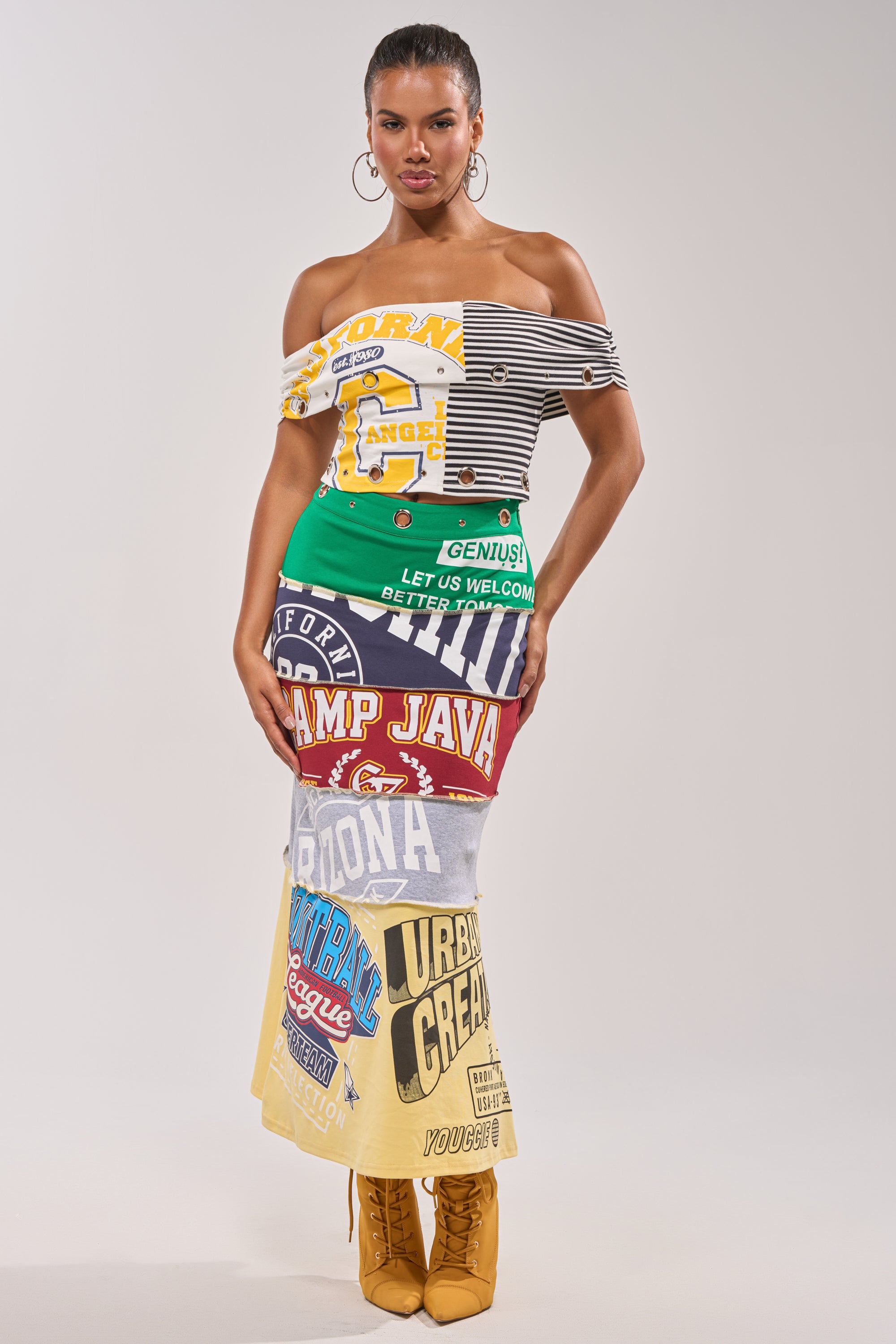 A woman poses confidently in a studio wearing the FULL RIDE PATCHWORK MAXI SKIRT, crafted from colorful graphic T-shirts, paired with an off-shoulder top and tan lace-up boots. She has slicked-back hair and neutral makeup against a plain background. 0