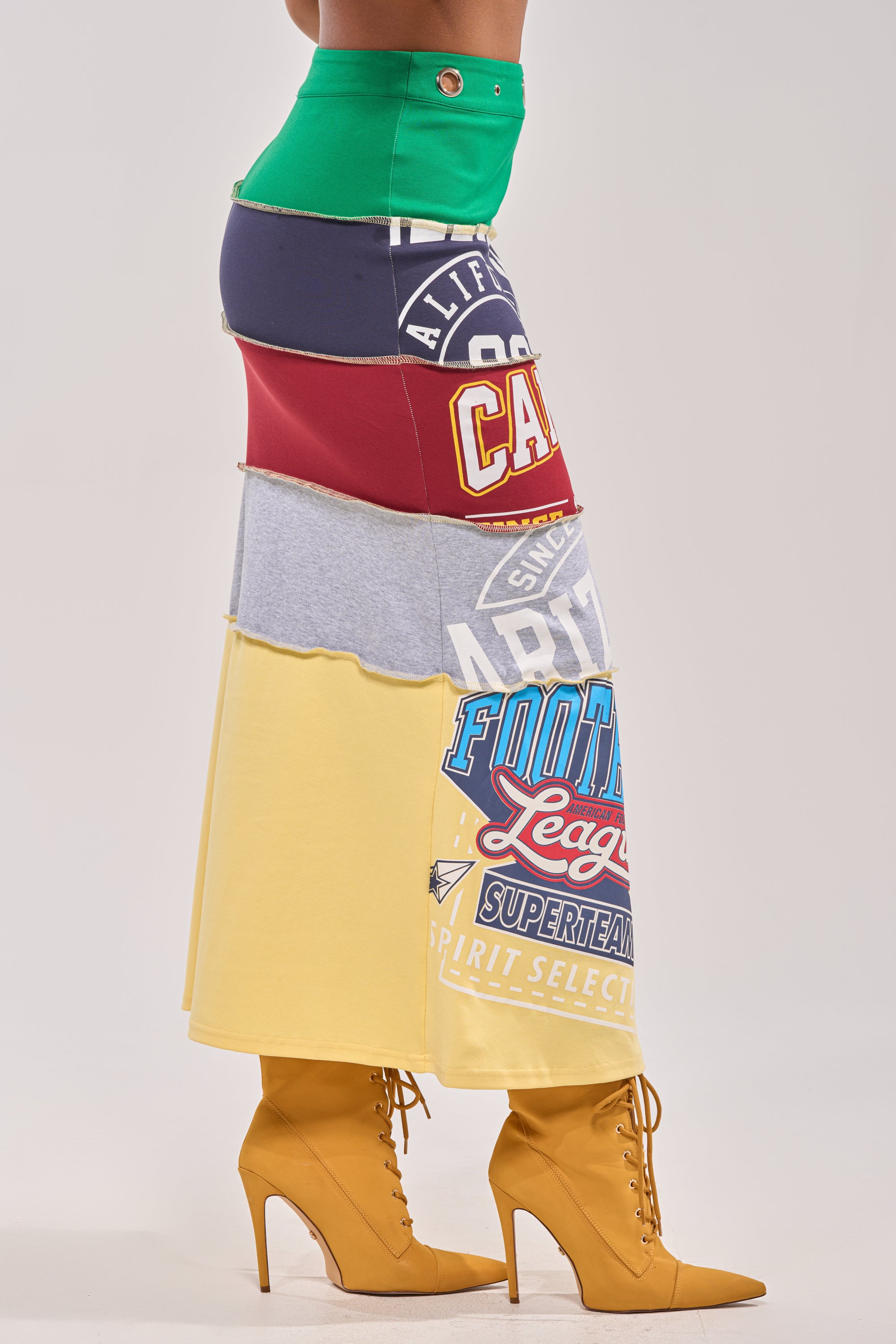A person wears the FULL RIDE PATCHWORK MAXI SKIRT, crafted from colorful repurposed graphic t-shirts, paired with mustard yellow high-heeled lace-up boots. The background is plain and light-colored. 3