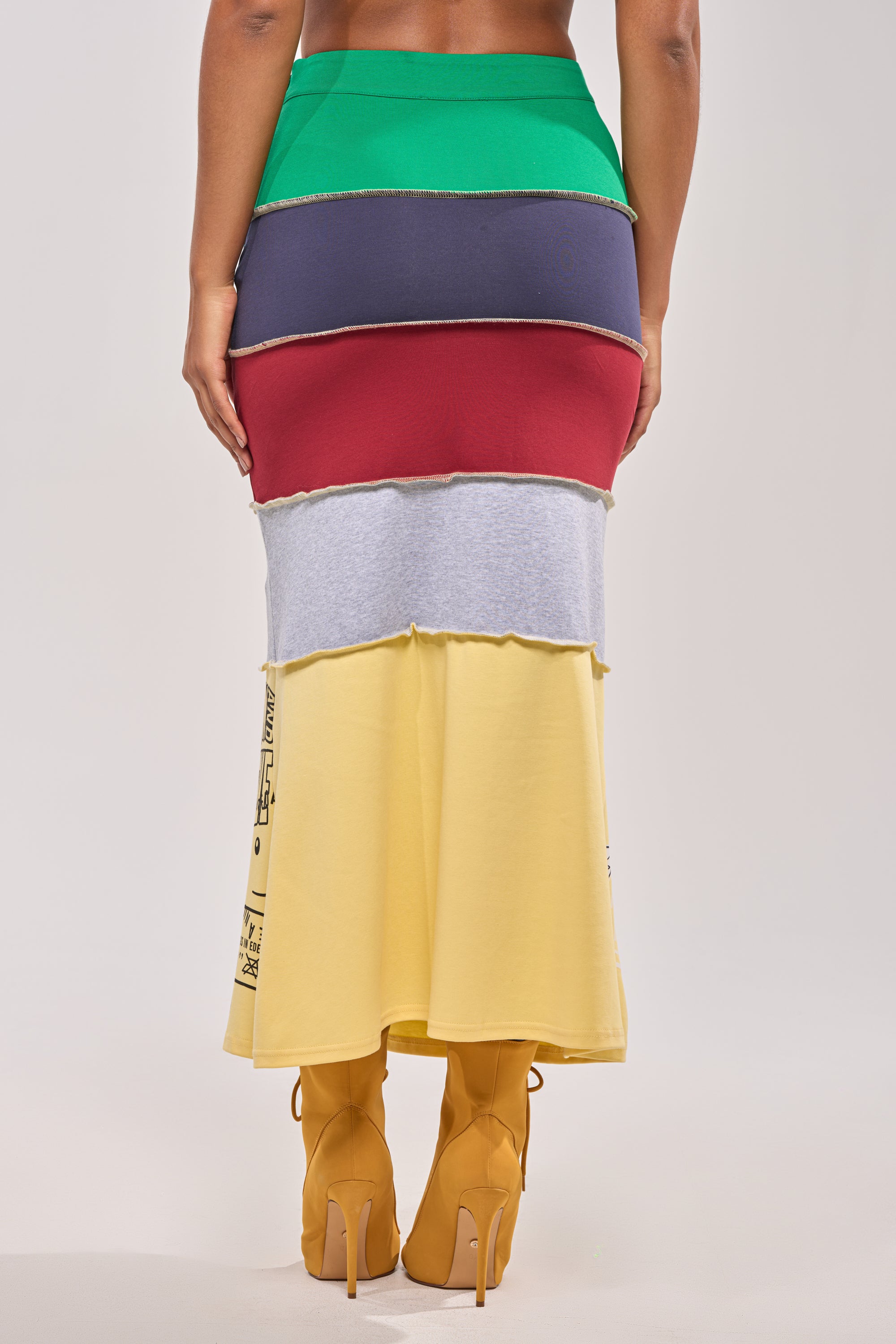 A person faces away, wearing the FULL RIDE PATCHWORK MAXI SKIRT with horizontal green, blue, red, gray, and yellow color blocks and yellow high-heeled boots against a plain light background. 4