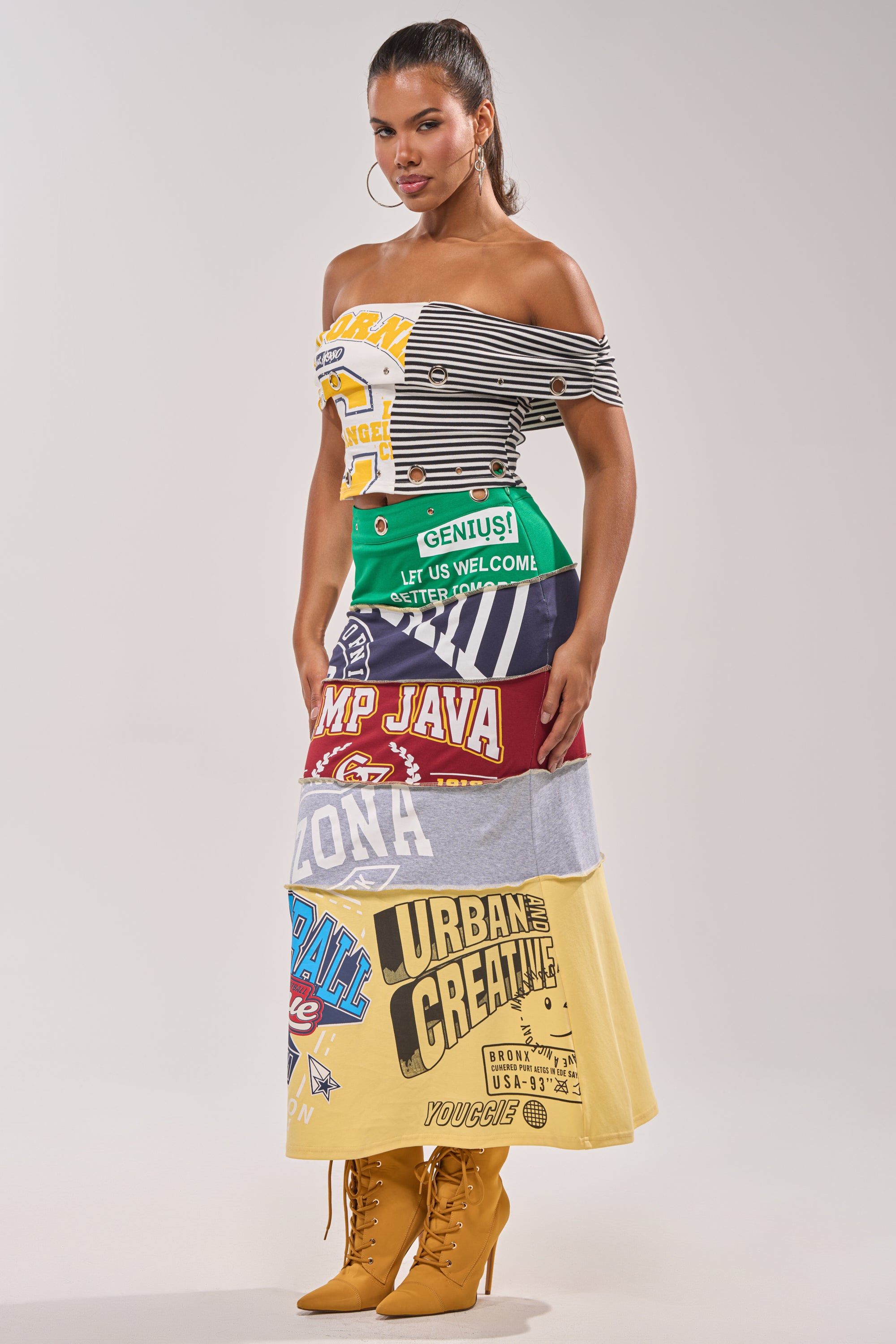 A woman showcases the FULL RIDE PATCHWORK MAXI SKIRT, made from repurposed graphic T-shirts with varsity patchwork details, paired with an off-shoulder striped top, tan lace-up boots, and hoop earrings for a courtside look. 5
