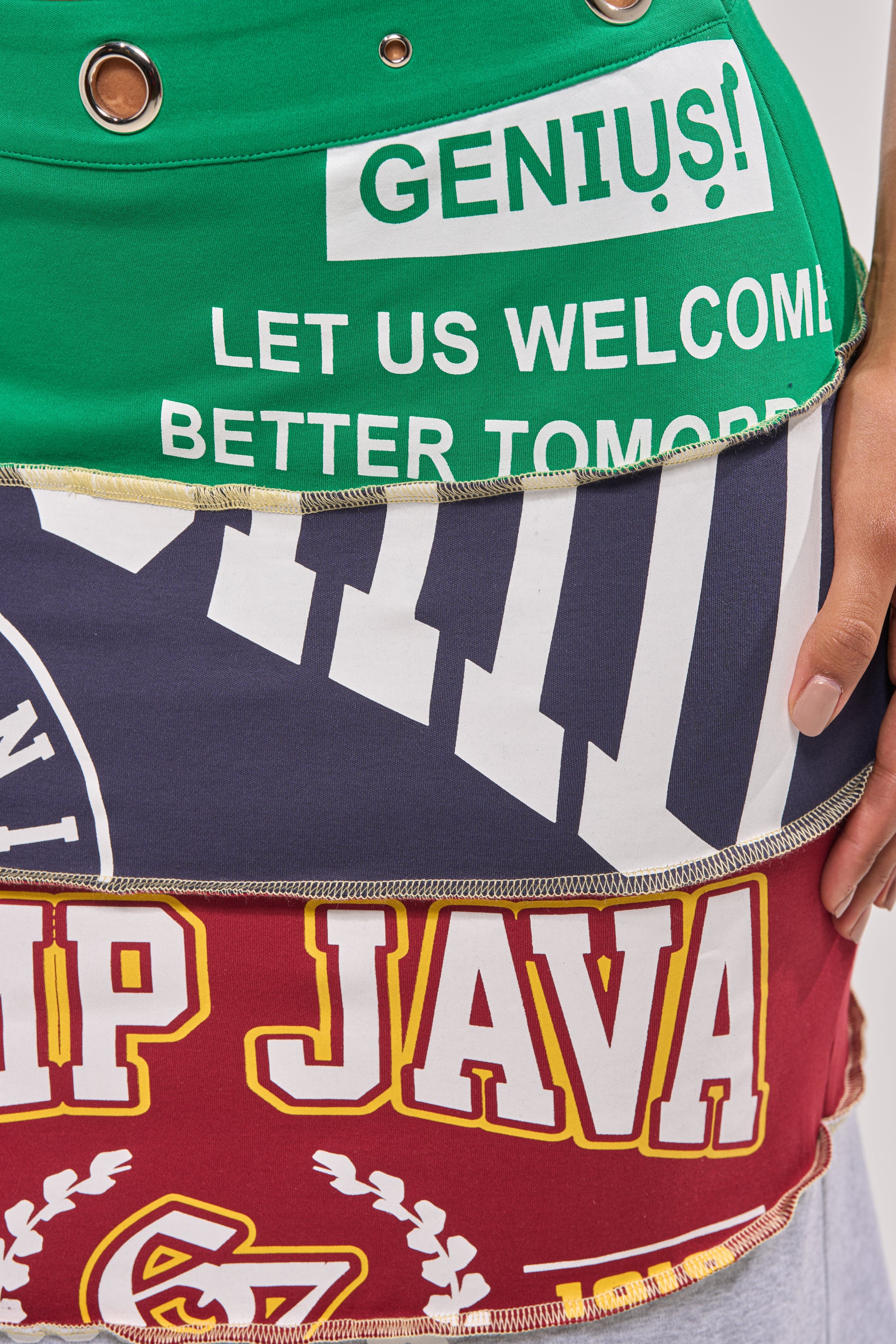 A close-up of the FULL RIDE PATCHWORK MAXI SKIRT, a varsity-inspired maxi made from stretchy cotton and layered, repurposed t-shirts in green, navy, and red with bold printed text like "GENIUS!" and "LET US WELCOME BETTER TOMOR. 7