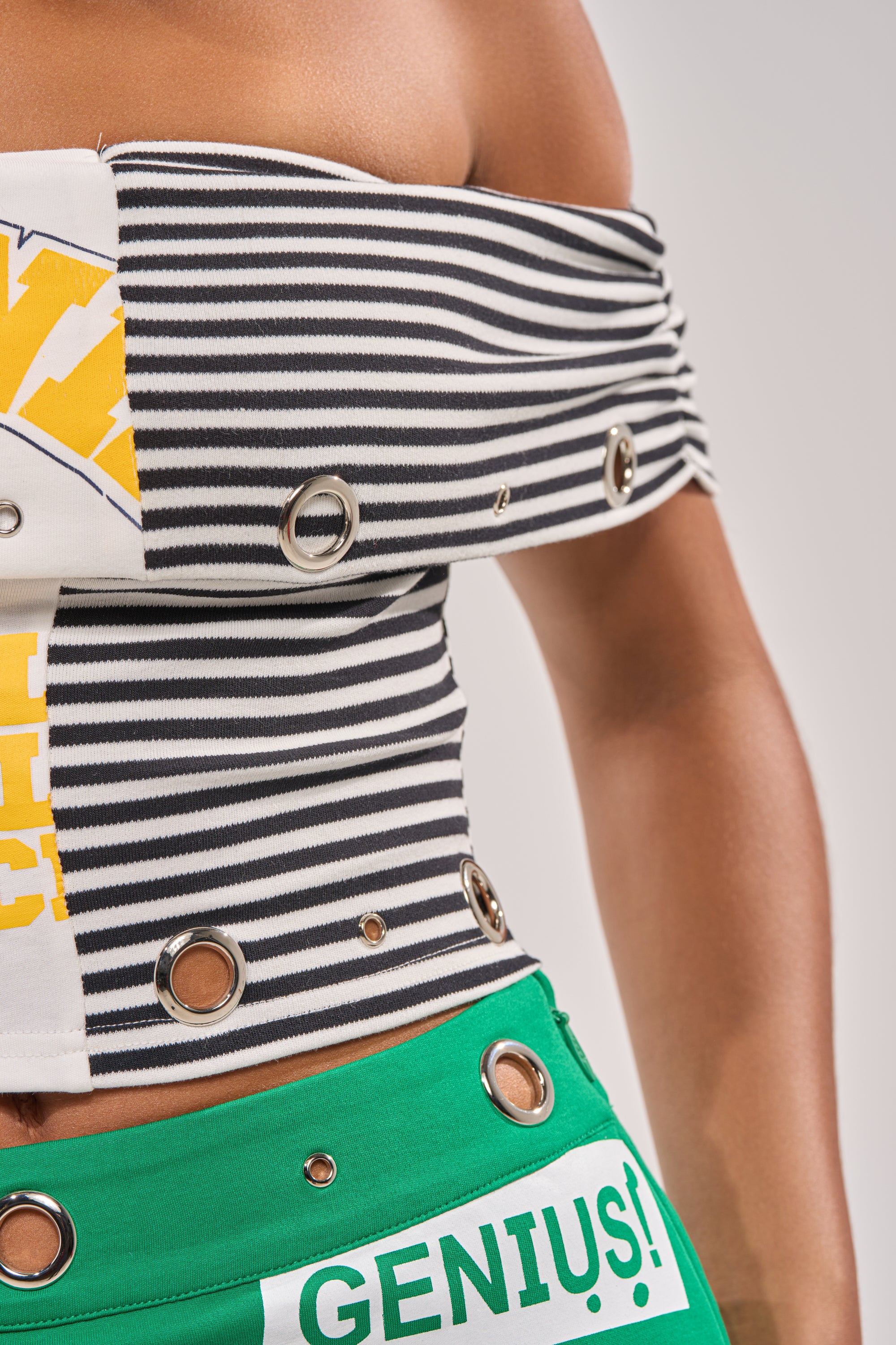 A close-up of a person in the FULL RIDE OFF THE SHOULDER PATCHWORK TOP, featuring black-and-white stripes and metal grommets, paired with green bottoms labeled "GENIUS!"—a striking streetwear look on a plain light background. 9