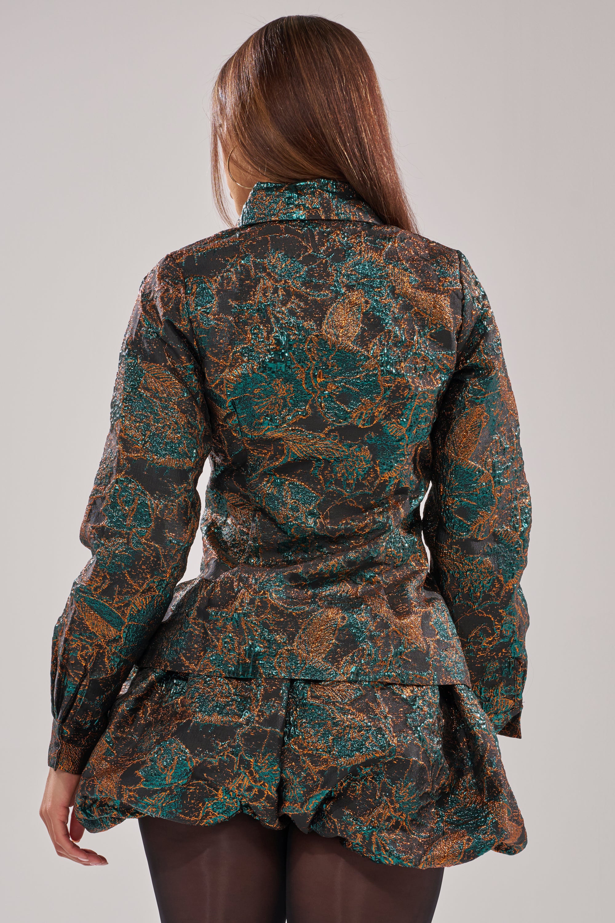 A woman with long brown hair stands facing away, wearing a dark AKIRA Label TOO HOT TO HANDLE BLOUSE with green and brown tones, long sleeves, and a ruffled hem. She pairs it with black tights and keeps her arms relaxed at her sides. 4
