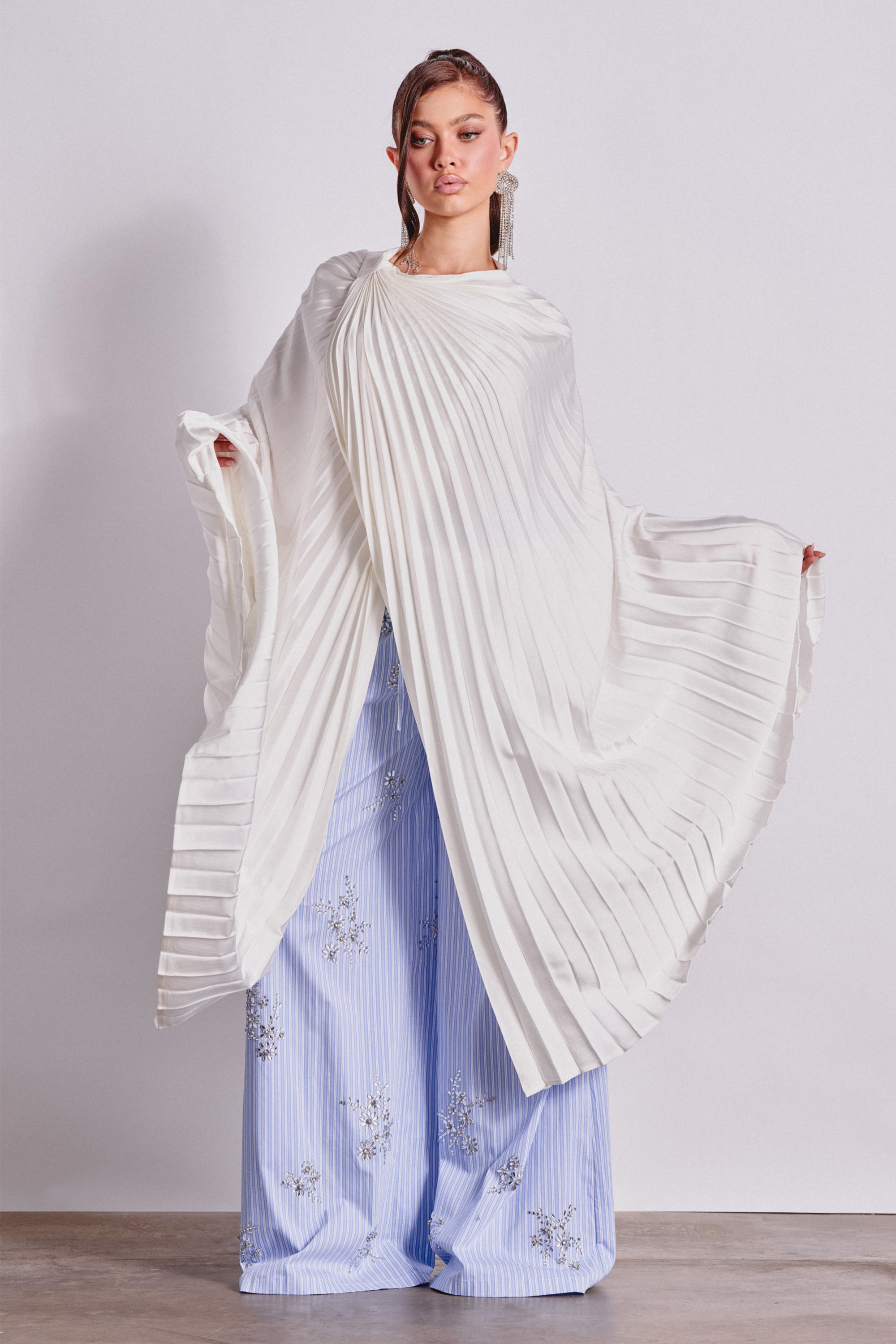 A person stands against a plain background, wearing the FARAH DRAPED PLEATED CAPE TOP IN WHITE over wide-legged, light blue pants with silver embroidery. They hold the cape open to display its sculptural pleats, embodying refined modern bridal style.