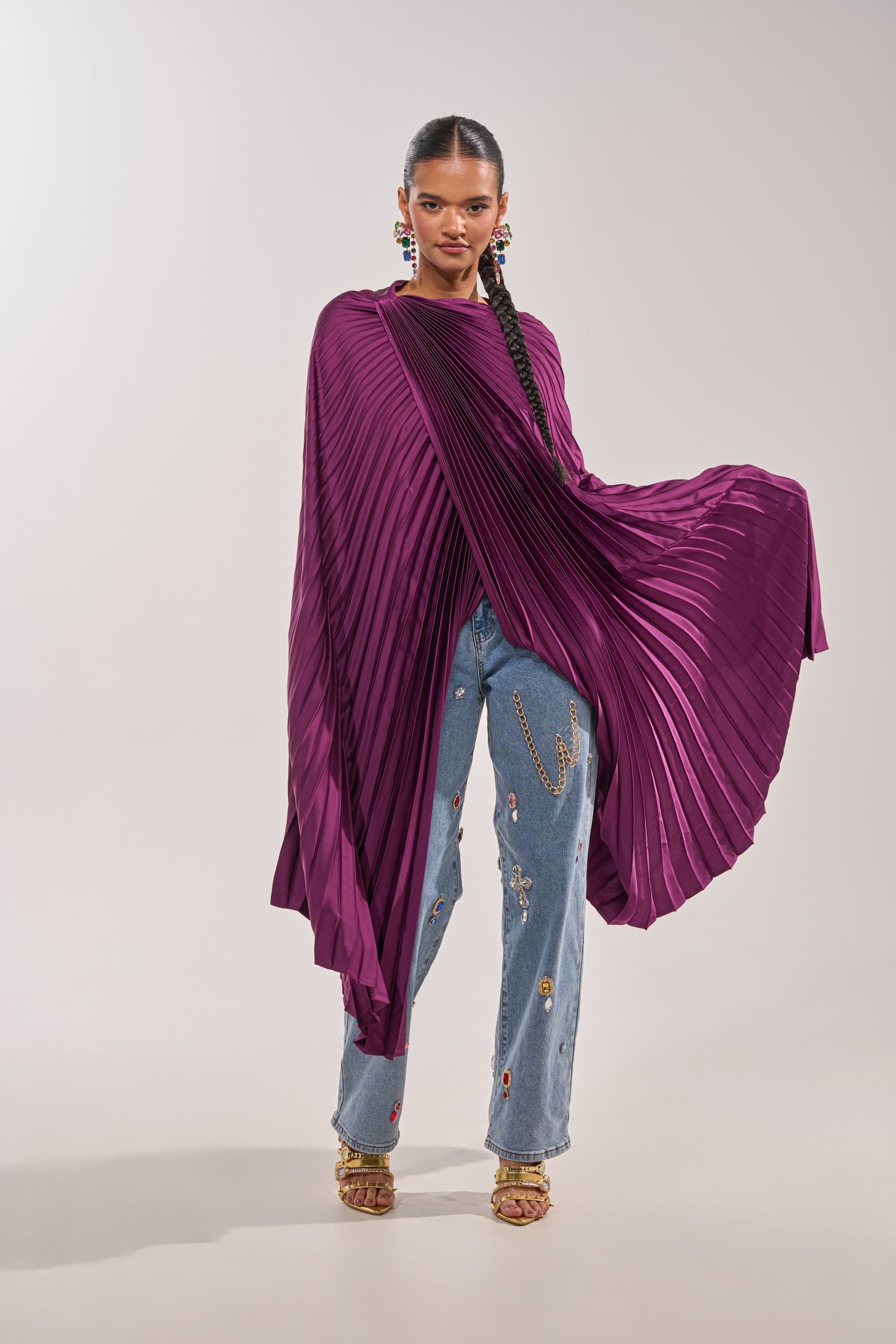 A woman exudes confidence in a runway-inspired look, sporting the FARAH DRAPED PLEATED CAPE TOP in deep purple with wide sleeves, styled with embellished light blue jeans and gold open-toe heels. 0