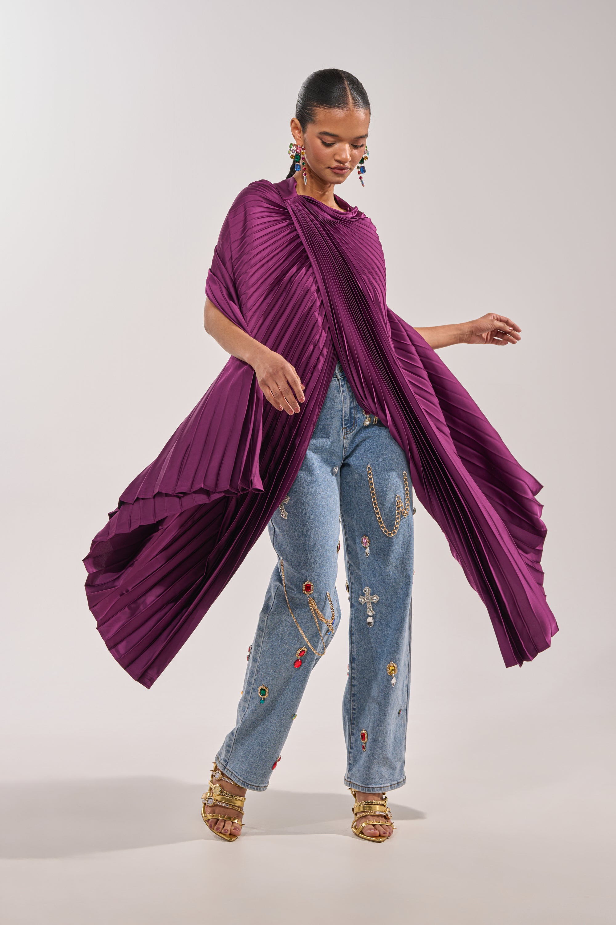 Wearing the FARAH DRAPED PLEATED CAPE TOP with light blue embroidered jeans, gold sandals, and statement earrings, a woman poses gracefully against a neutral background—a chic look inspired by runway fashion. 1