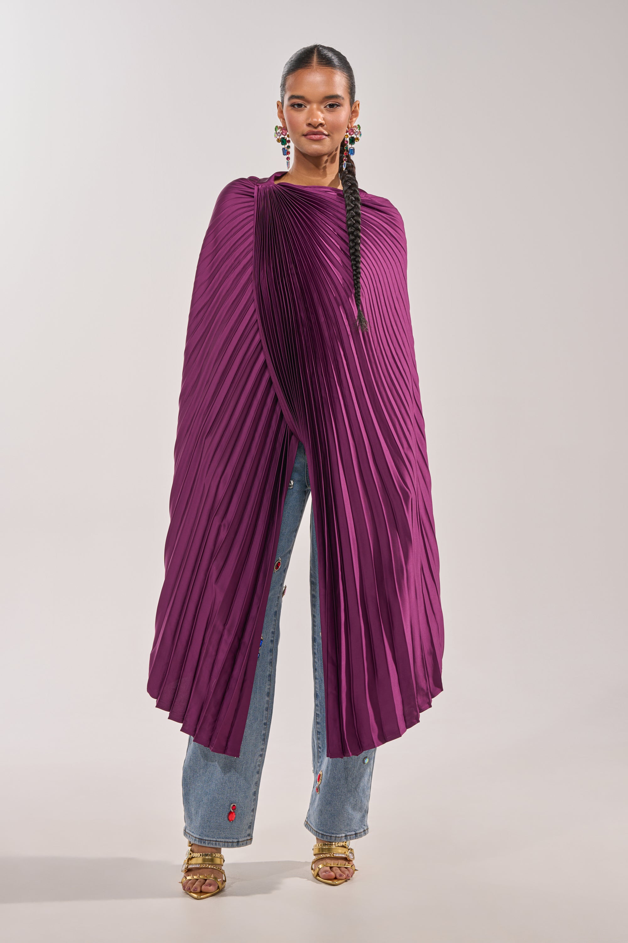 A woman wears the FARAH DRAPED PLEATED CAPE TOP in vibrant purple over embellished blue jeans and gold heels. Her braided hair and colorful statement earrings complete the sculptural evening look against a plain light background. 2