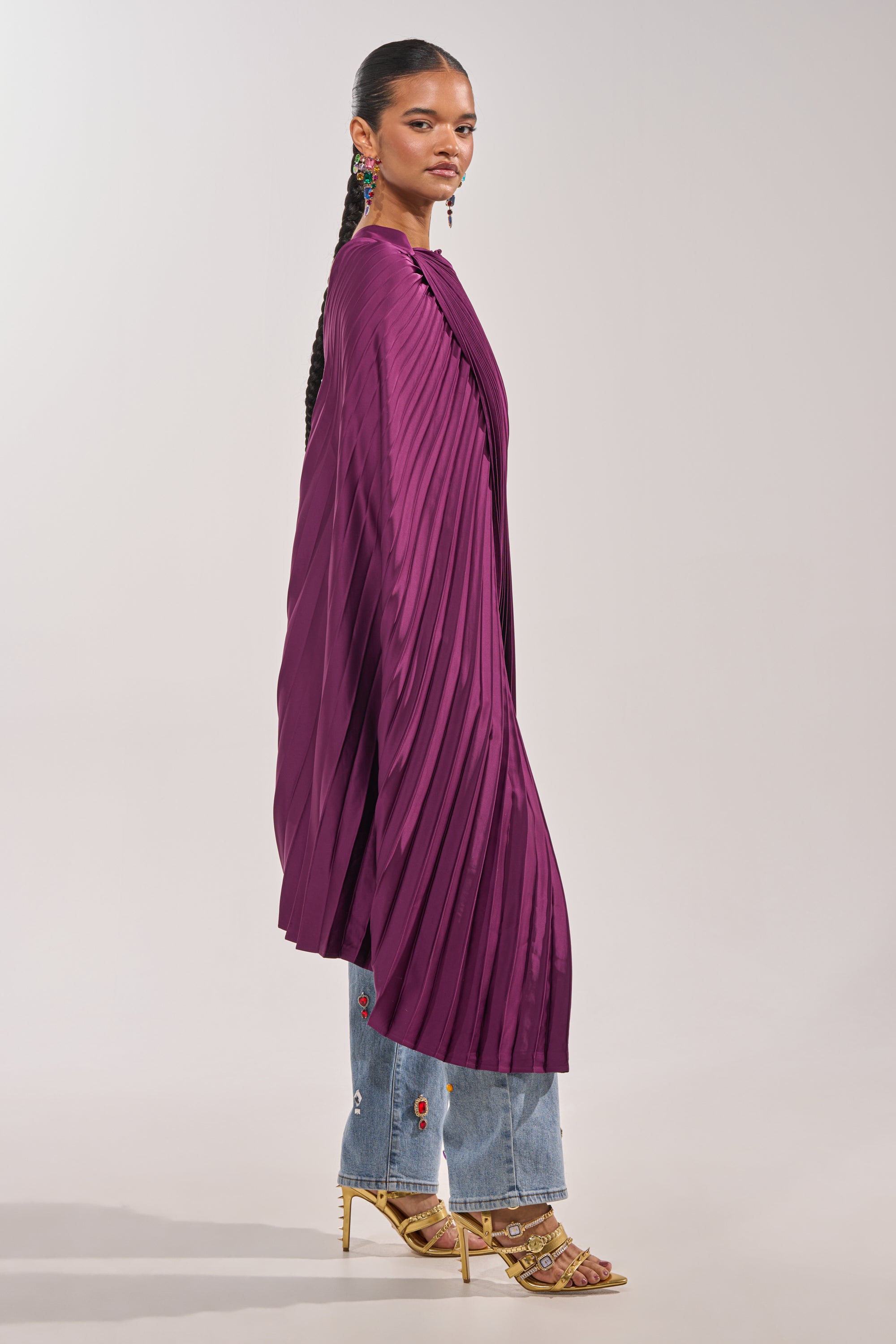 A person stands sideways against a plain light gray background, wearing the FARAH DRAPED PLEATED CAPE TOP in purple over light blue floral-embroidered jeans and yellow strappy heels for a striking evening look. 3