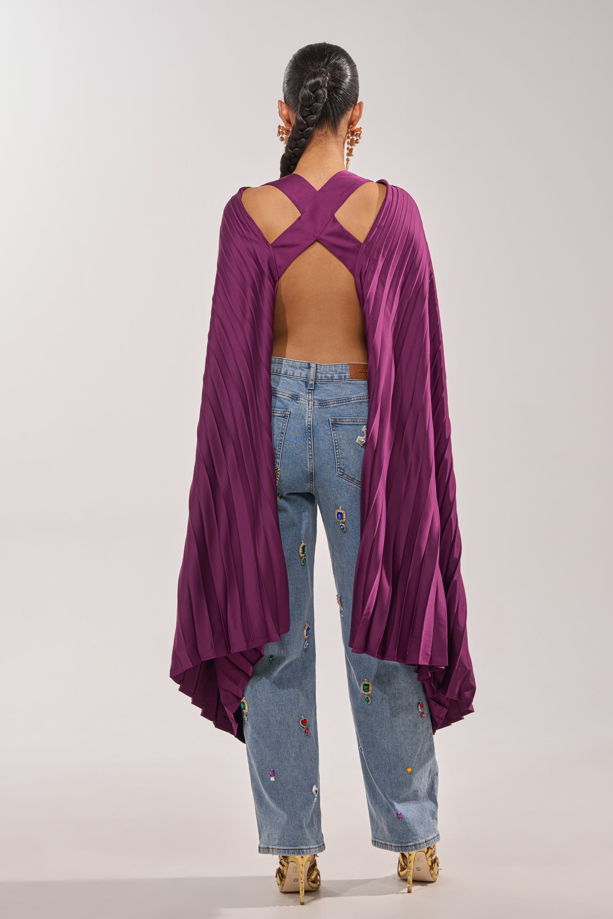 A person stands facing away in the FARAH DRAPED PLEATED CAPE TOP with purple pleated sleeves, a crisscross open-back, light blue embroidered jeans, gold heels, and a long braided hairstyle. 4