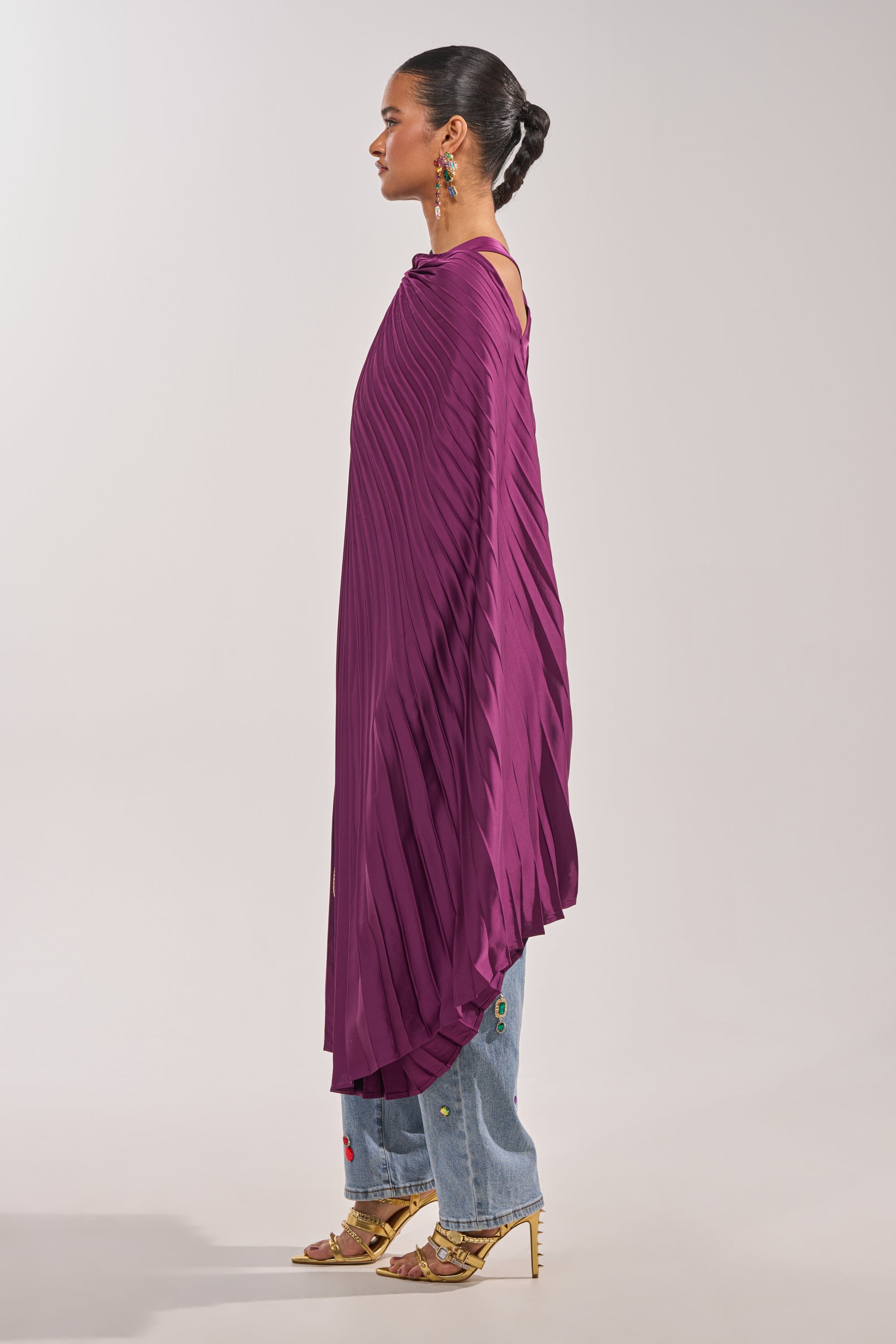 A woman stands in profile against a light gray background, wearing the FARAH DRAPED PLEATED CAPE TOP in purple over embroidered blue jeans, gold high-heeled sandals, slicked-back hair, and long dangling earrings. 5