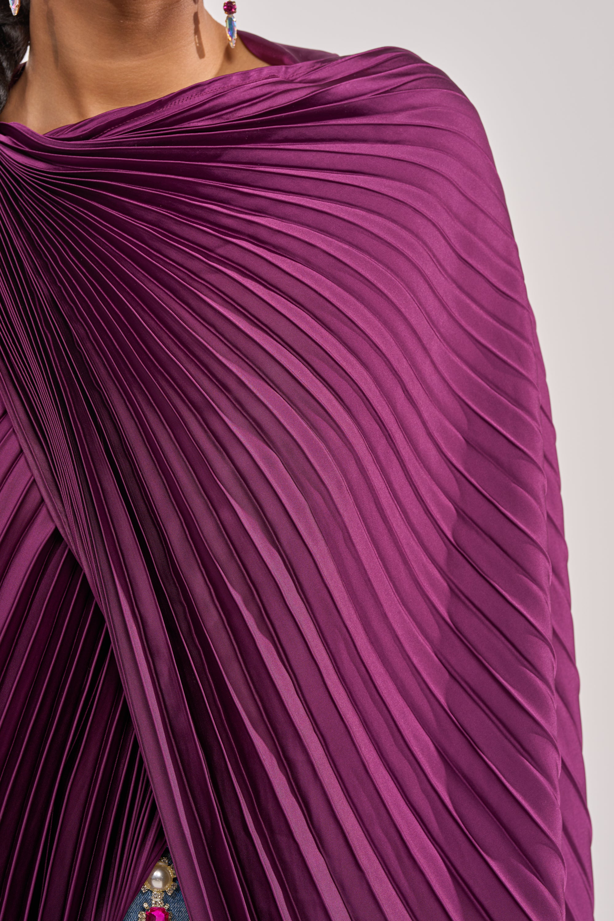 A close-up shows a person wearing the FARAH DRAPED PLEATED CAPE TOP in purple, elegantly draped over the shoulder with part of an earring and their neck visible—perfect for statement evening wear. 6