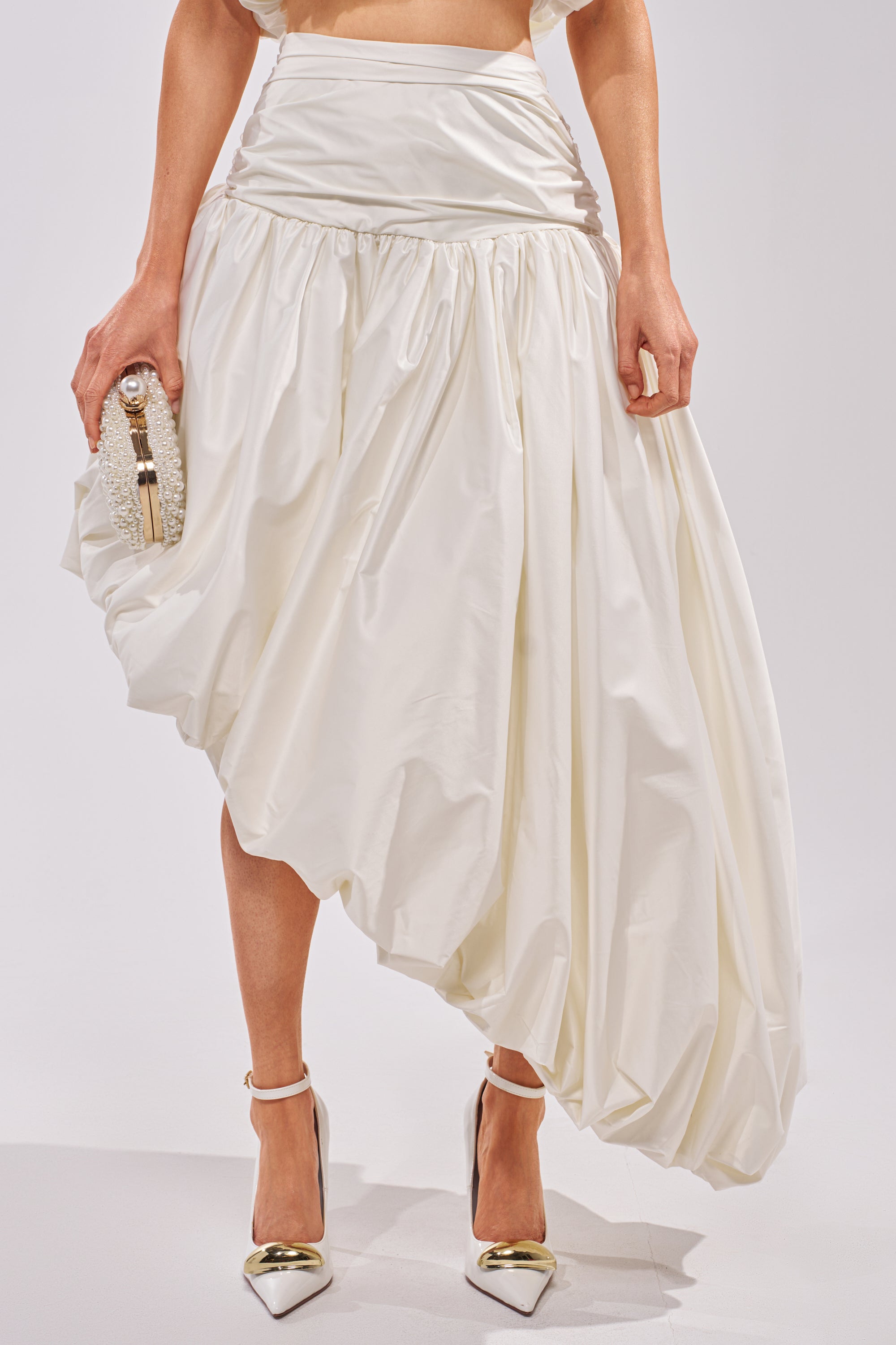 Wearing the HEAD IN THE CLOUDS HIGH LOW SKIRT in off-white with a ruched waist and asymmetrical hem, paired with white high heels and a small textured clutch, a person stands against a plain background. 1