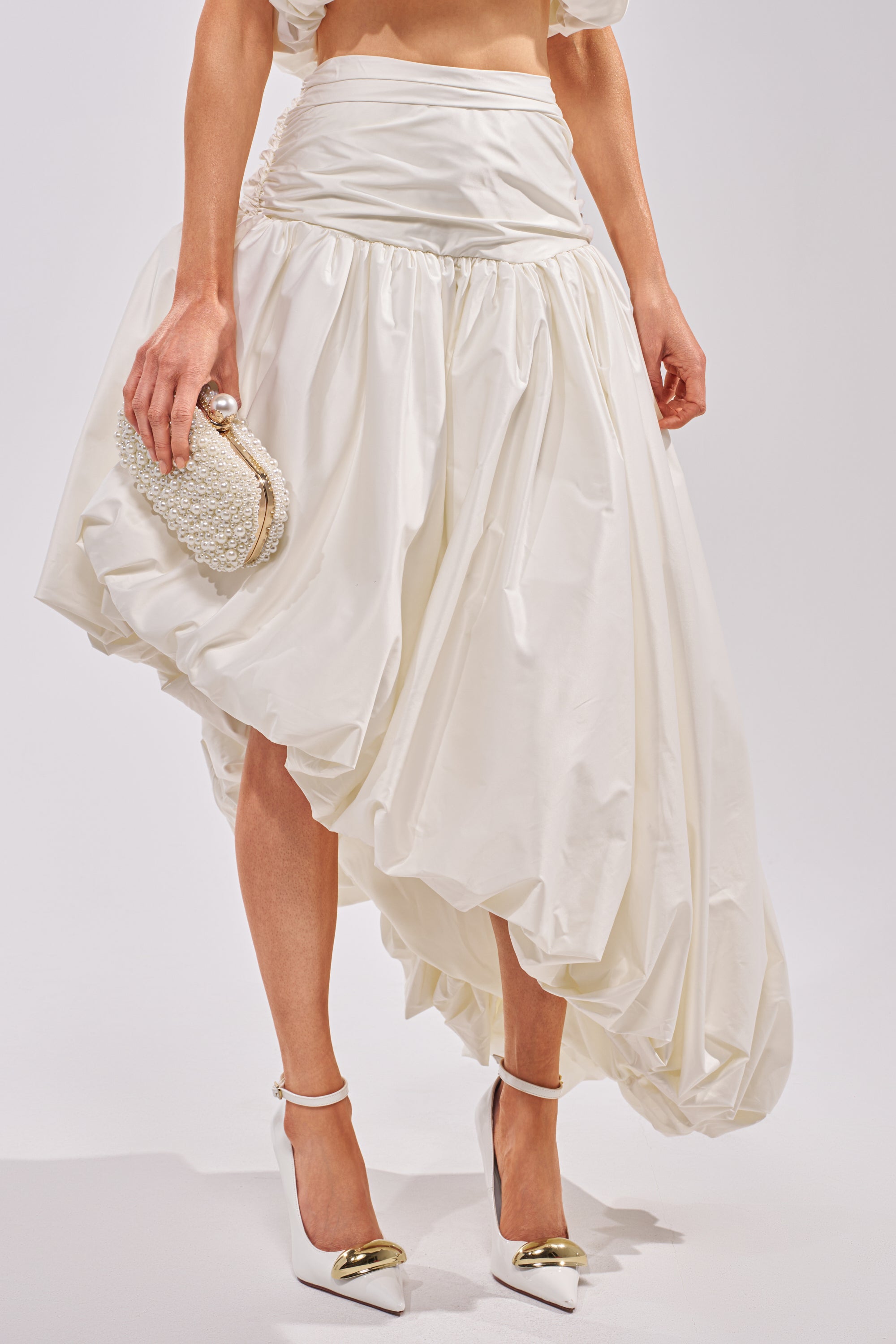 A woman wears the HEAD IN THE CLOUDS HIGH LOW SKIRT, a white asymmetrical high-low design, with white high heels featuring gold-capped toes and holds a small textured white clutch against a plain background. 2