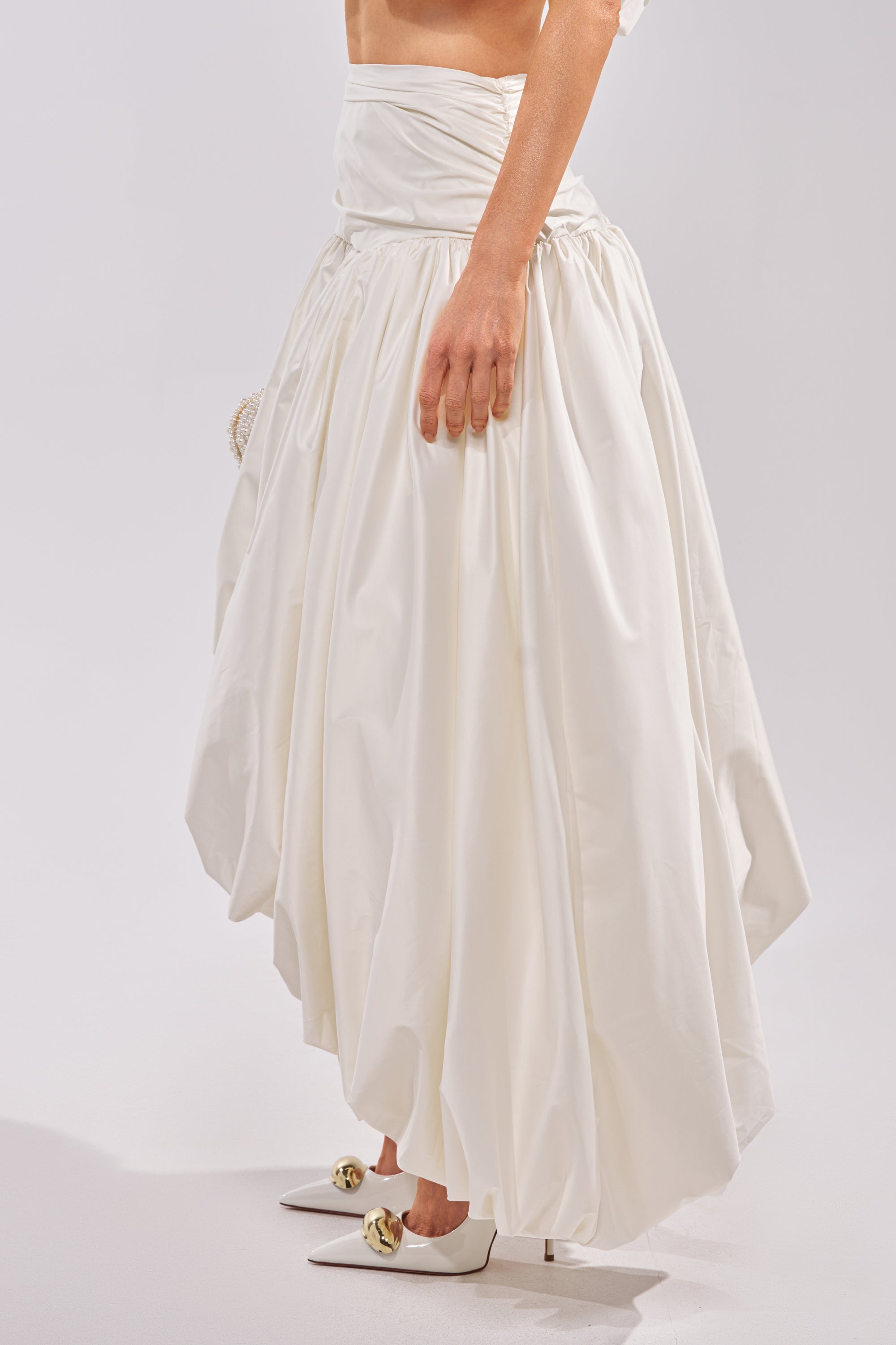 Wearing the HEAD IN THE CLOUDS HIGH LOW SKIRT and white pointed-toe heels with gold accents, a person stands against a plain light background, showing only their lower torso and legs. 3