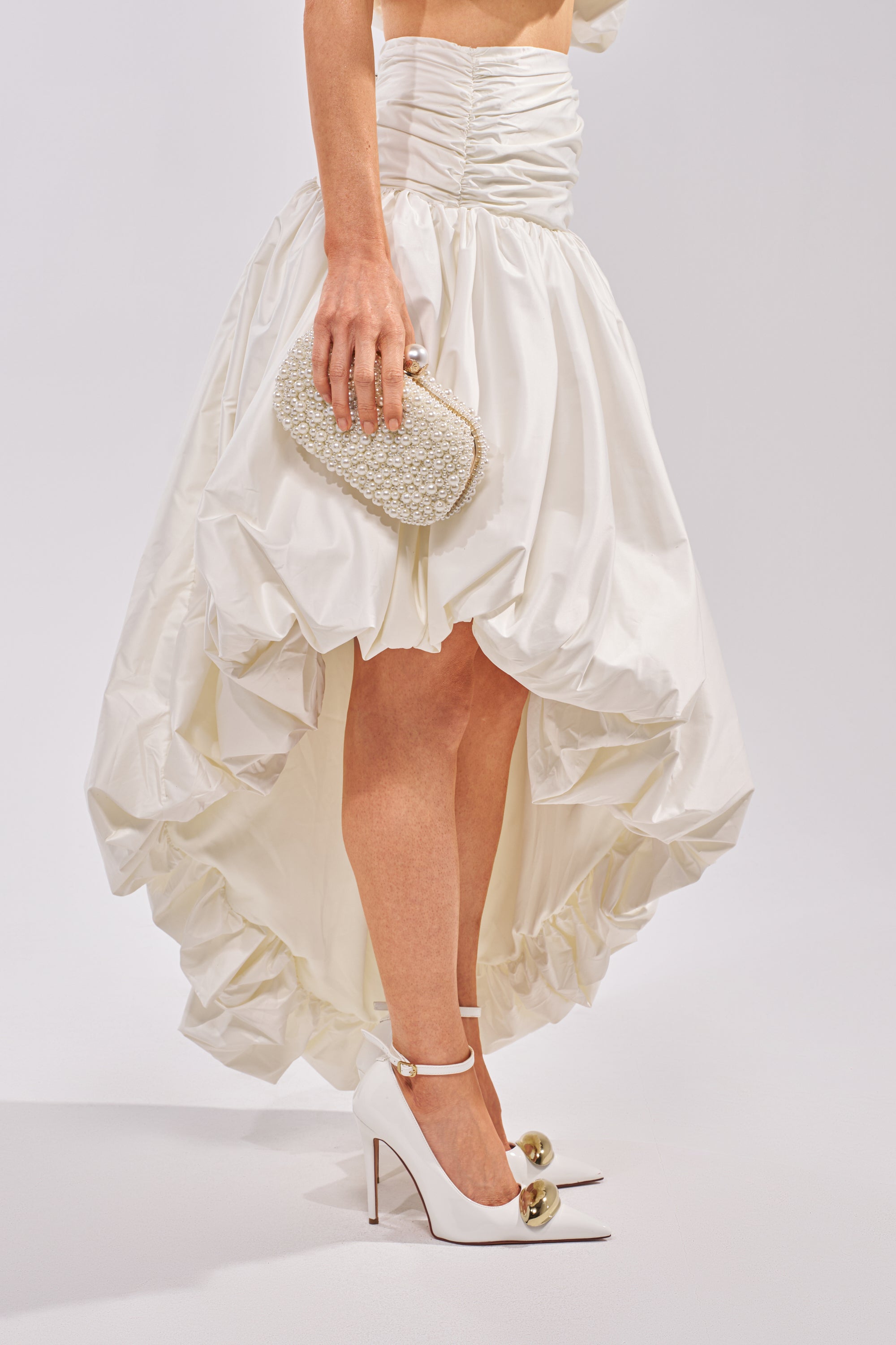 A person wears the HEAD IN THE CLOUDS HIGH LOW SKIRT, white high heels with gold ball details, and holds a small textured white clutch against a plain background. 4