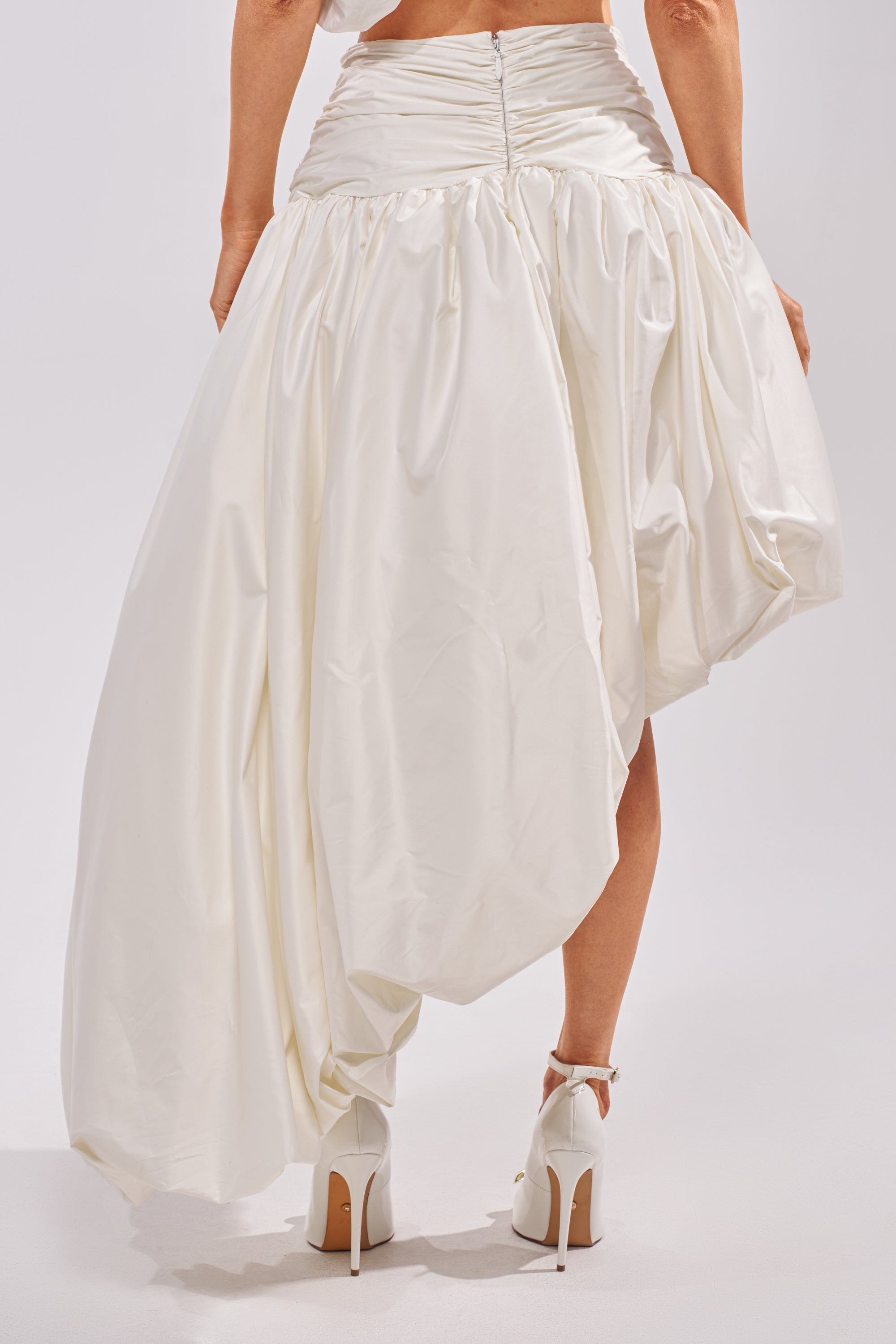 Someone is photographed from the back against a plain background, wearing the HEAD IN THE CLOUDS HIGH LOW SKIRT—an asymmetrical, voluminous white satin skirt with ruching at the waist—paired with high-heeled white shoes. 5