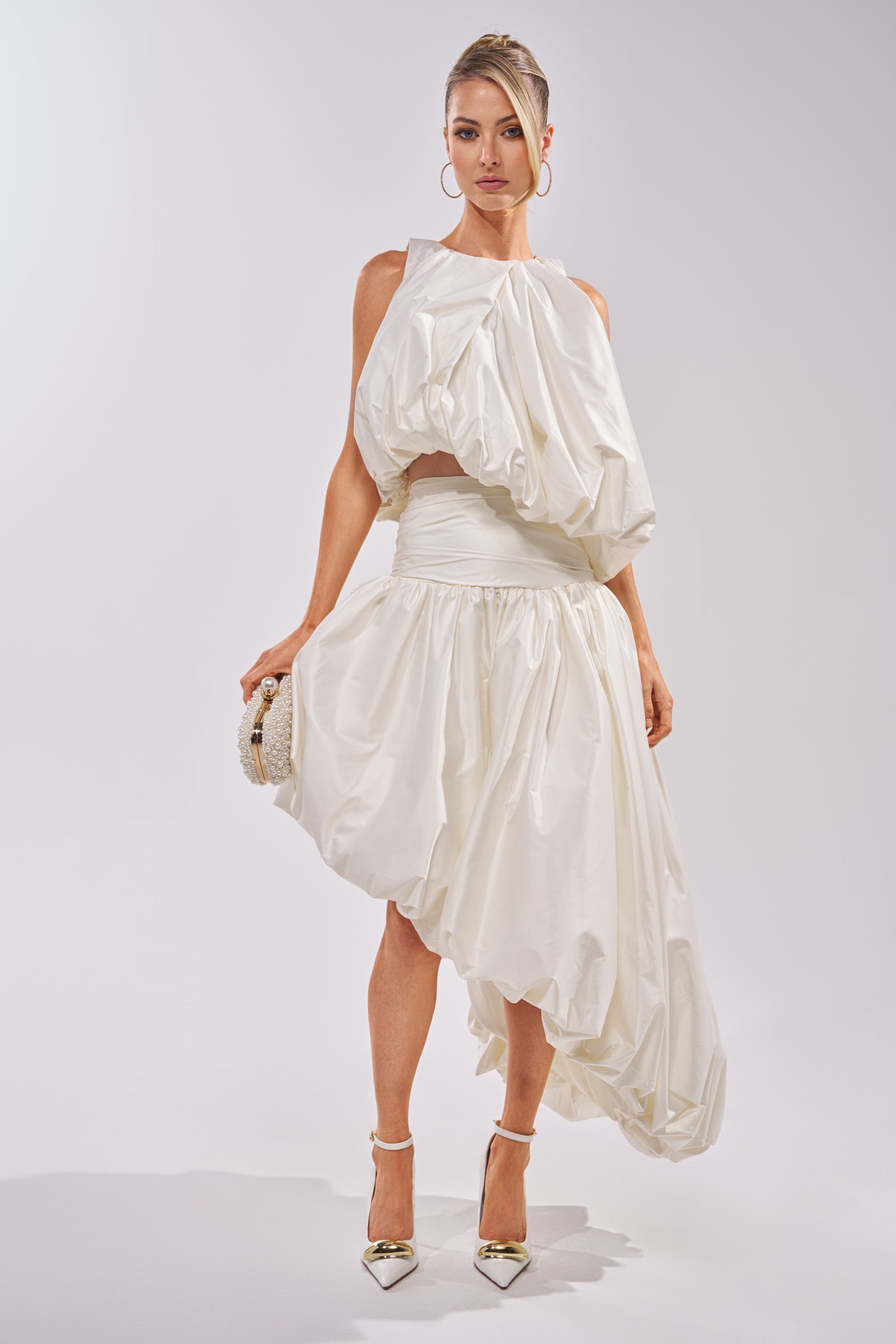 A woman poses against a plain background wearing the HEAD IN THE CLOUDS HIGH LOW SKIRT, featuring voluminous white fabric with an asymmetrical hemline. She accessorizes with a small textured clutch, hoop earrings, and white pointed heels. 6