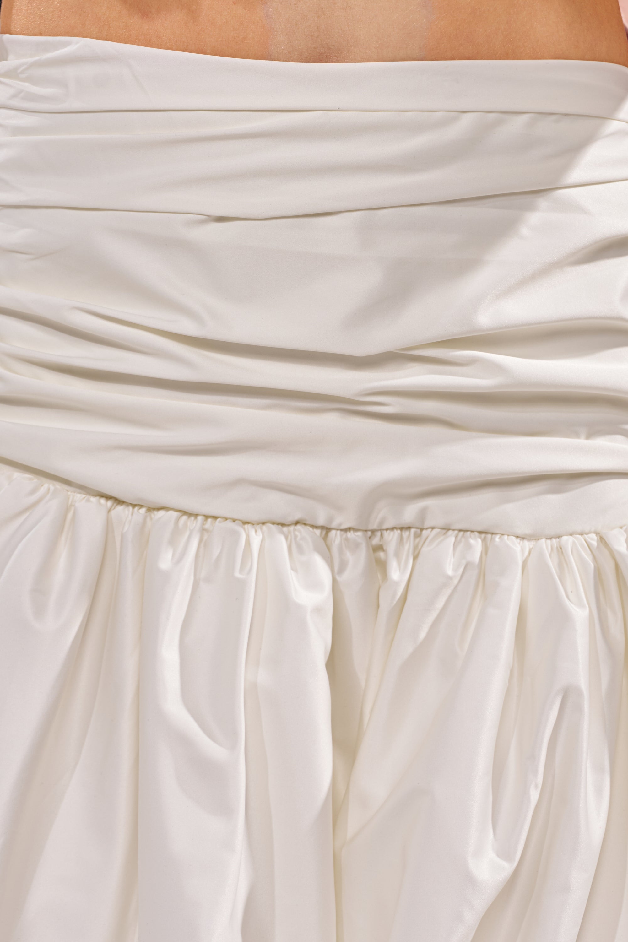 Close-up of the HEAD IN THE CLOUDS HIGH LOW SKIRT, featuring cream satin fabric, a ruched waistband, and an asymmetrical high-low hemline. The image highlights the skirt's unique design and luxurious sheen. 7