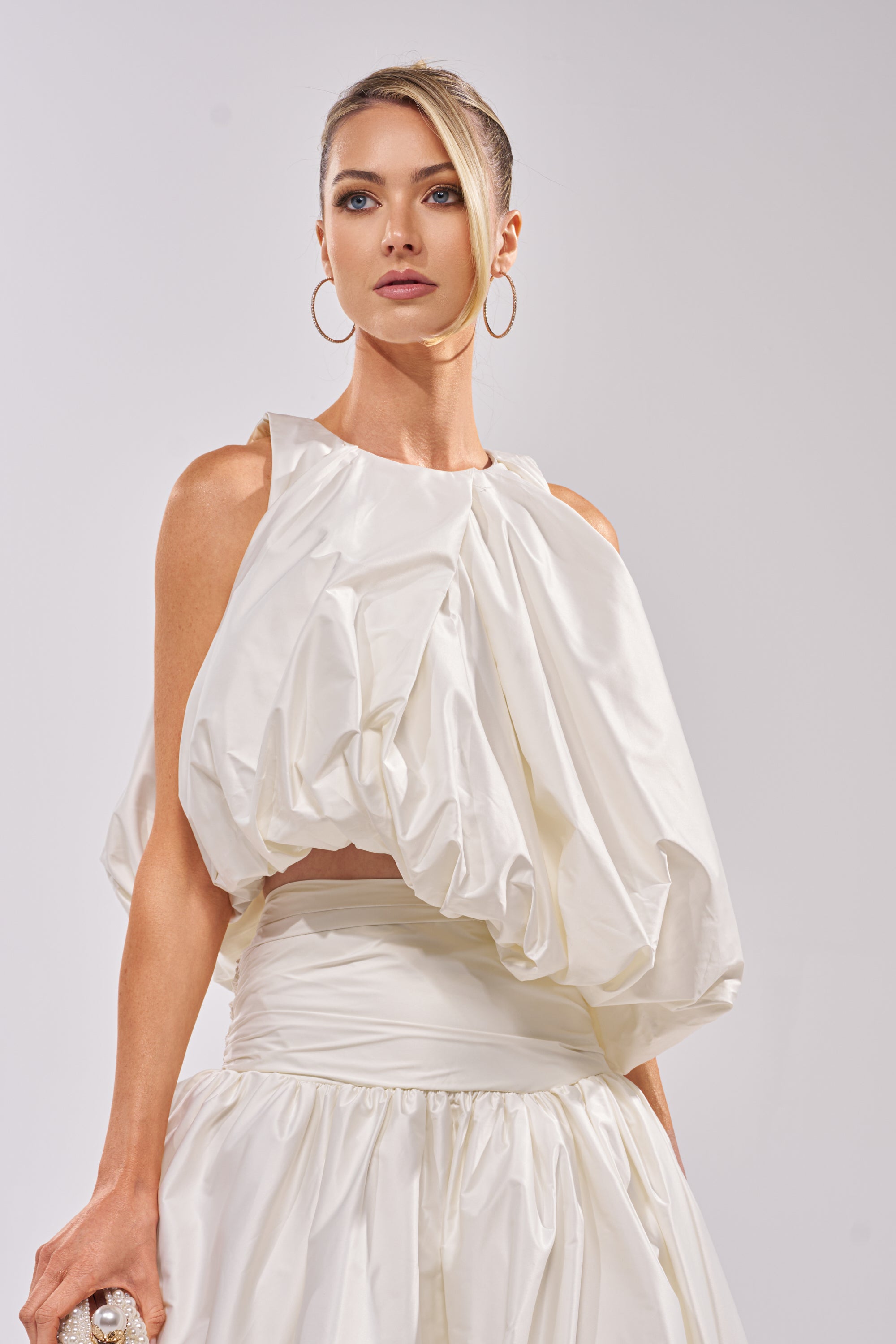 A woman stands confidently against a plain light background, wearing the HEAD IN THE CLOUDS SLEEVELESS BLOUSE with a voluminous bubble silhouette and gathered, draped top, paired with hoop earrings. 0