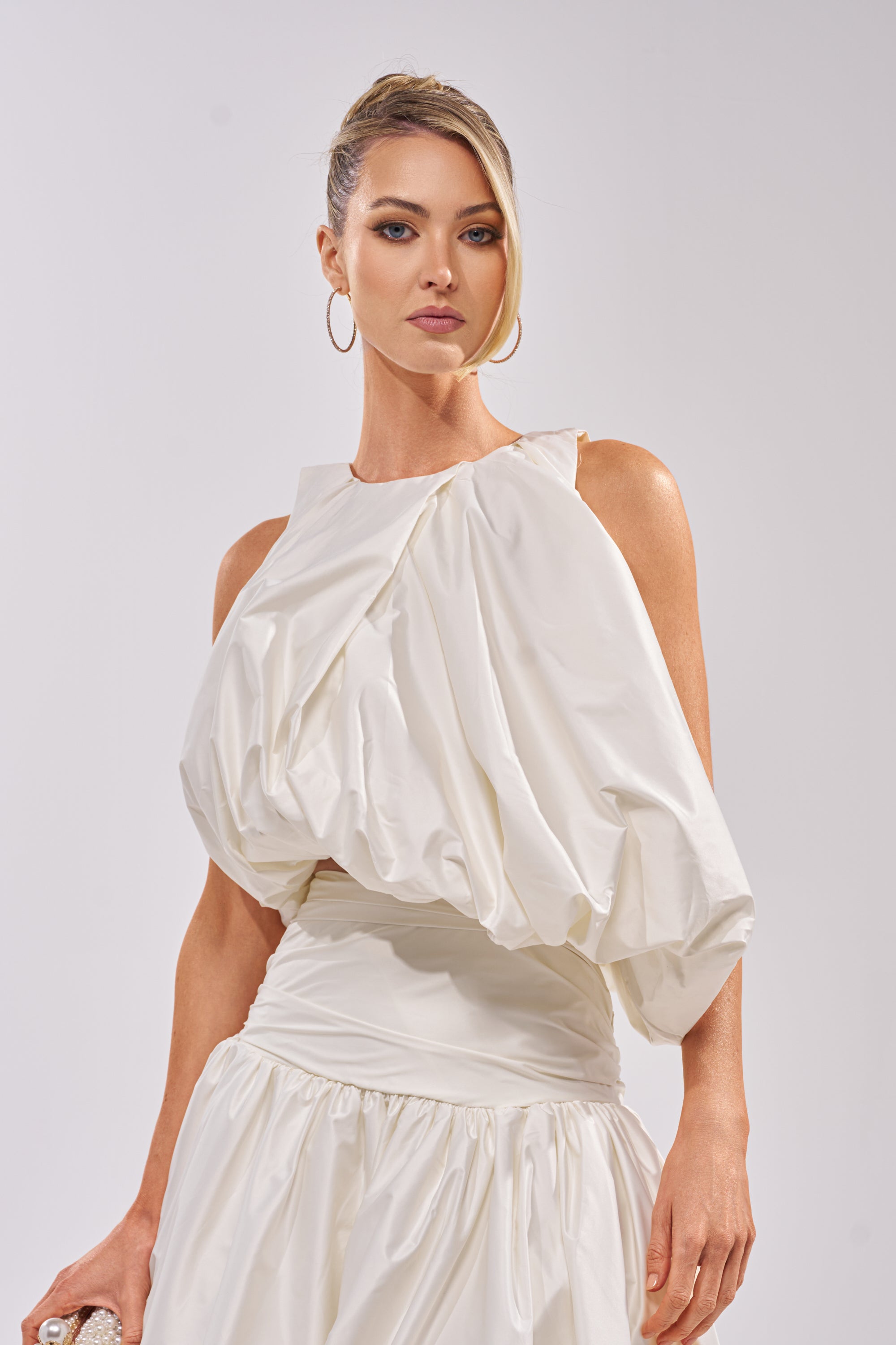 A woman with blonde hair in an elegant updo wears the HEAD IN THE CLOUDS SLEEVELESS BLOUSE, a modern white top with a bubble silhouette and gathered bodice, posing confidently against a plain light background. 1