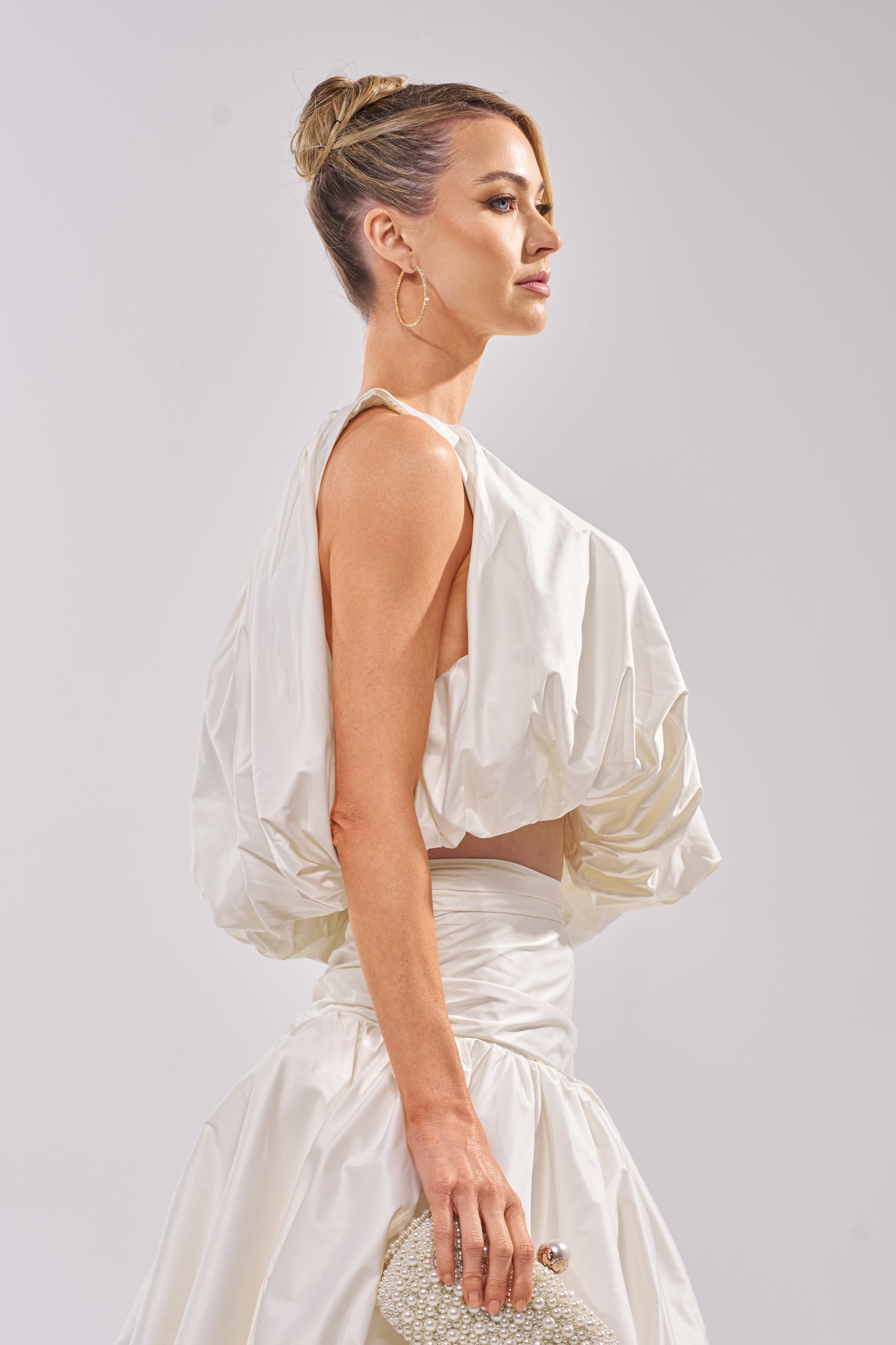 A woman with blonde hair in a neat bun wears the HEAD IN THE CLOUDS SLEEVELESS BLOUSE, a stylish white, voluminous top with a bubble silhouette, and holds a beaded clutch while standing in profile against a light gray background. 2