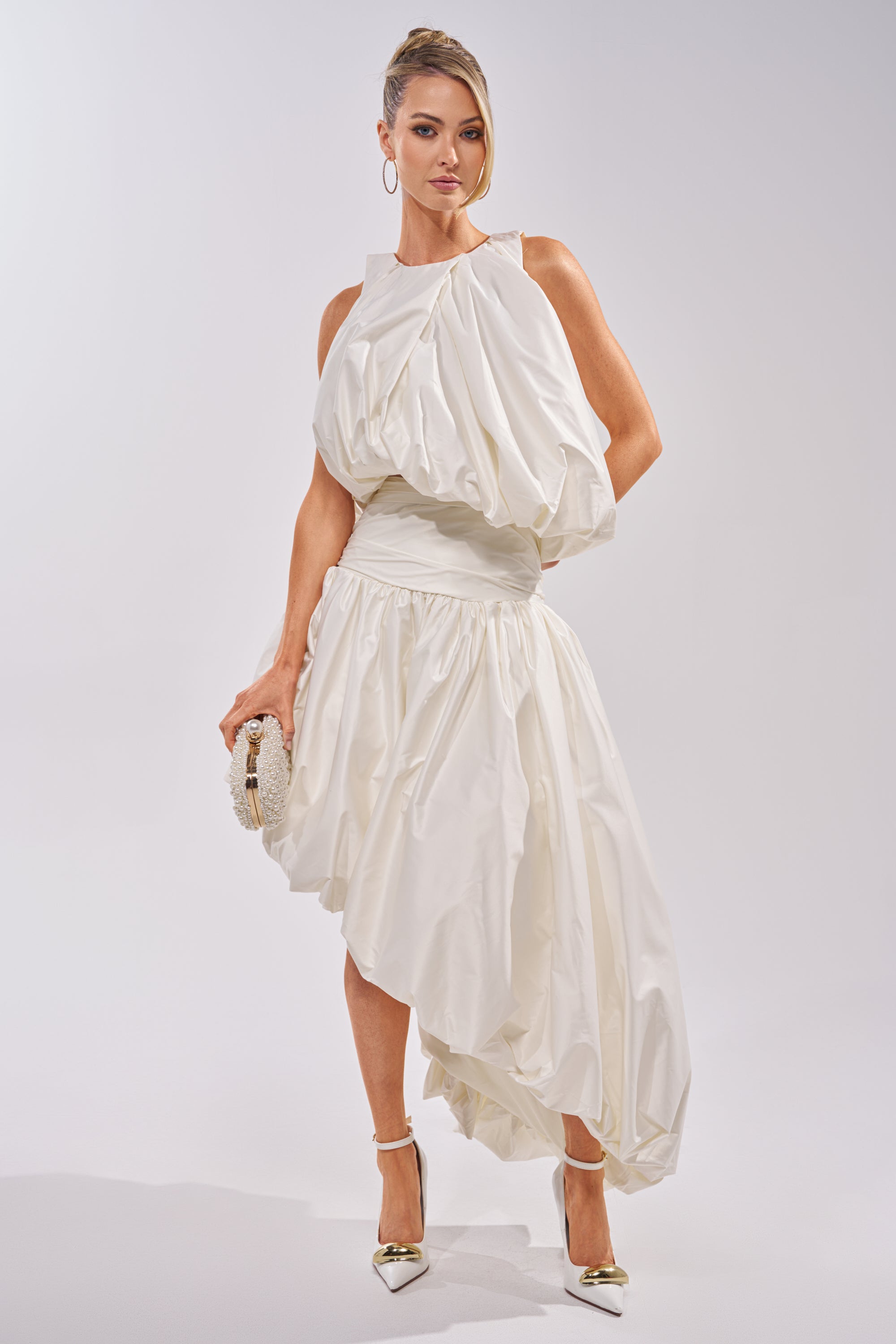 A woman poses against a plain light background wearing the HEAD IN THE CLOUDS SLEEVELESS BLOUSE, styled with pointed heels, hoop earrings, a small clutch, and her hair up. 5