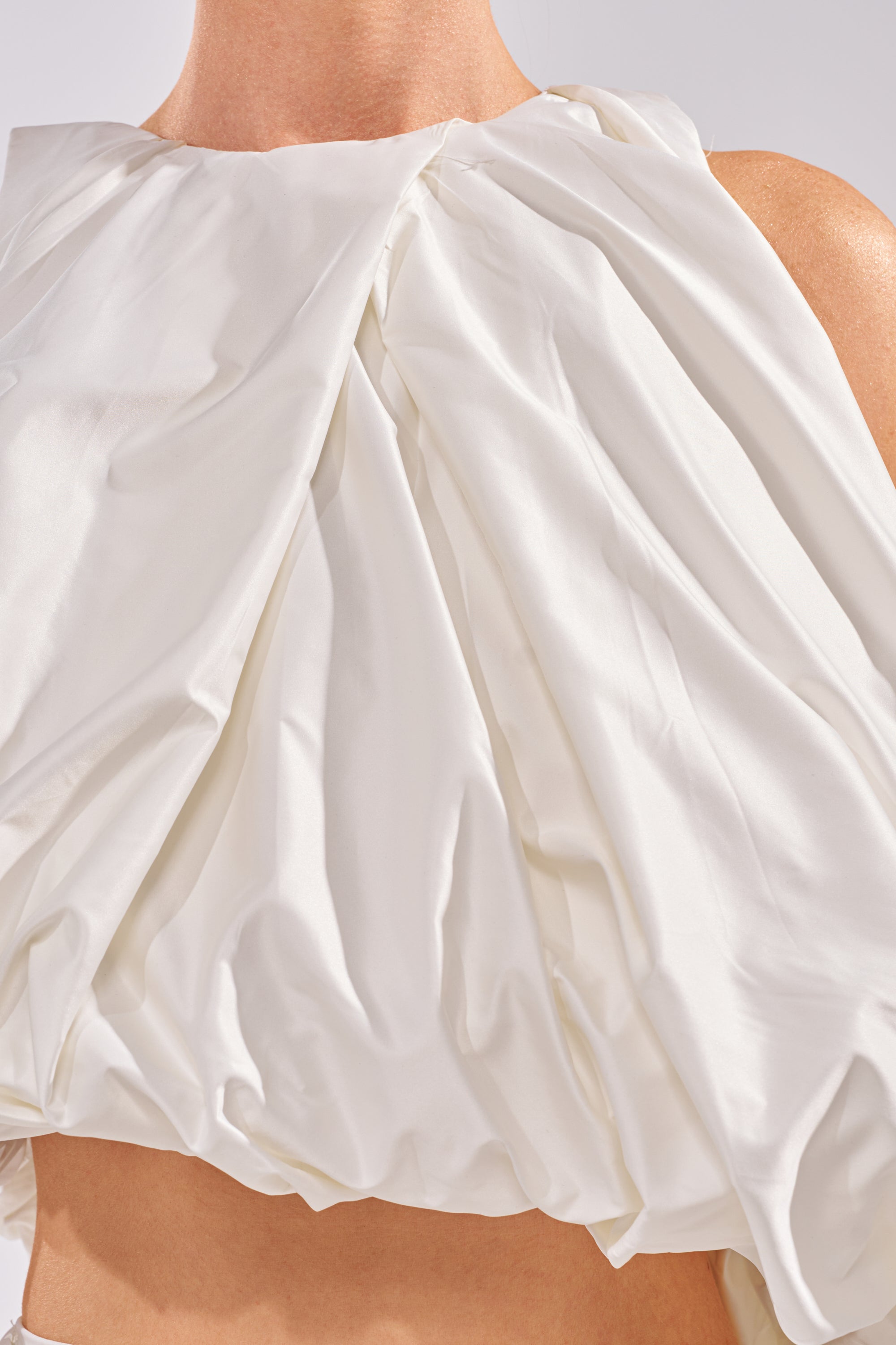 A close-up of a person wearing the HEAD IN THE CLOUDS SLEEVELESS BLOUSE, featuring glossy white satin with draped and ruched details. The photo is cropped just below the shoulders and above the waist to showcase its unique texture. 6