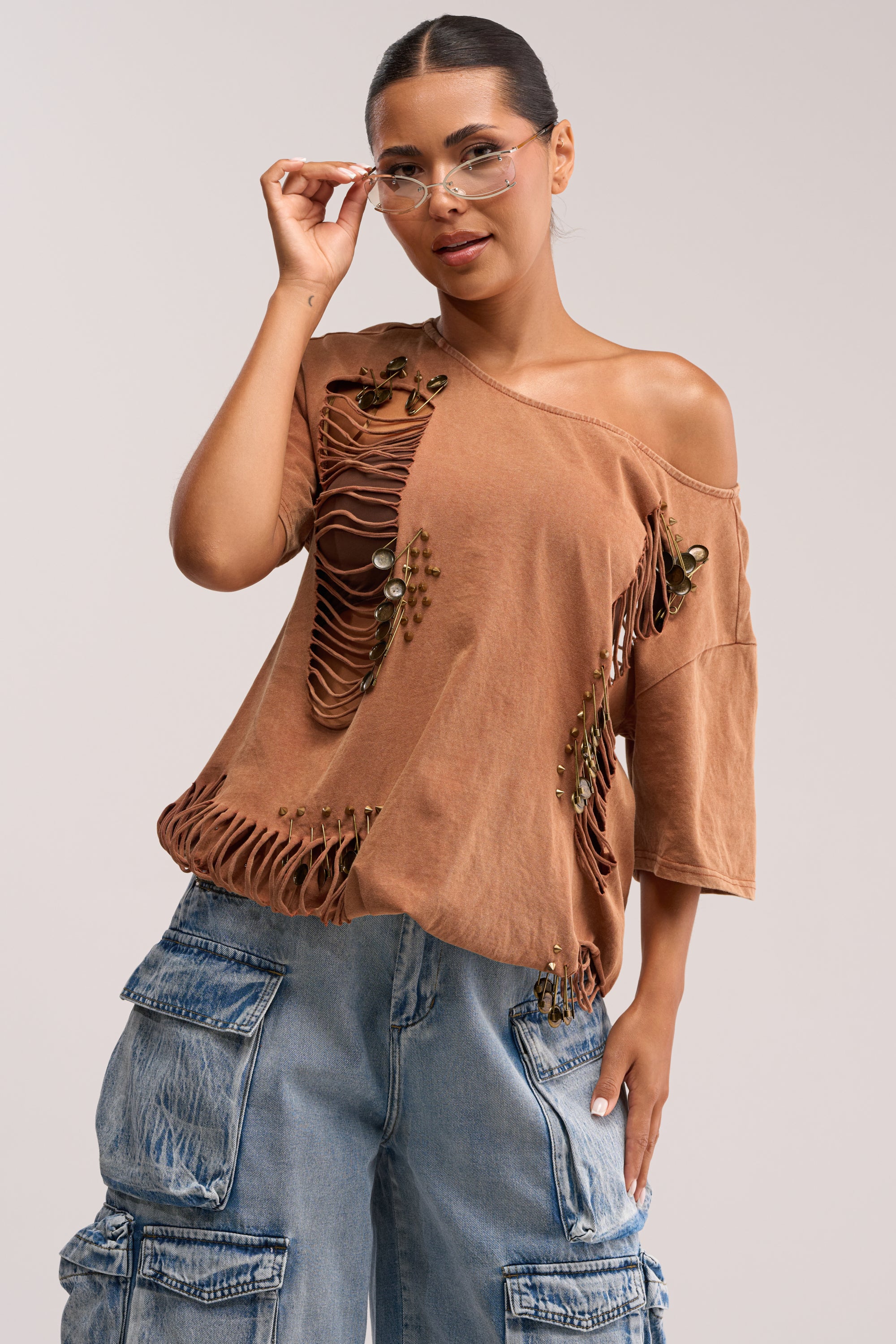 A woman wears the IN THE CUT DISTRESSED TEE with shredded paneling and cutout details, teamed with clear glasses and loose blue cargo jeans, posing confidently against a neutral background.