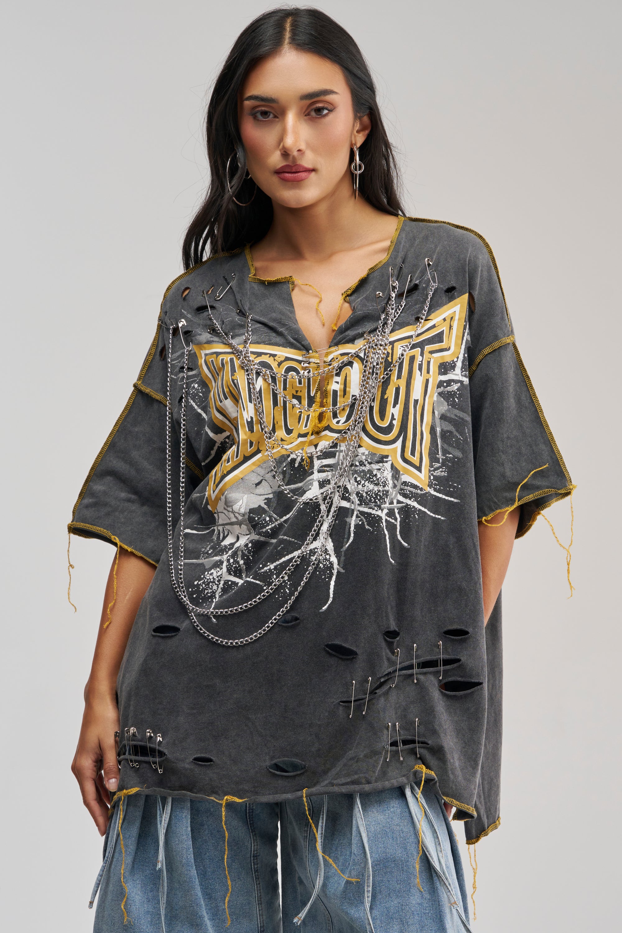 A woman with long dark hair wears the KNOCKOUT OVERSIZED TEE IN GREY MULTI, featuring distressed details, yellow stitching, metal chains, and ripped accents—an edgy streetwear staple. She pairs it with faded blue jeans and stands against a plain background, effortlessly embodying standout festival style.