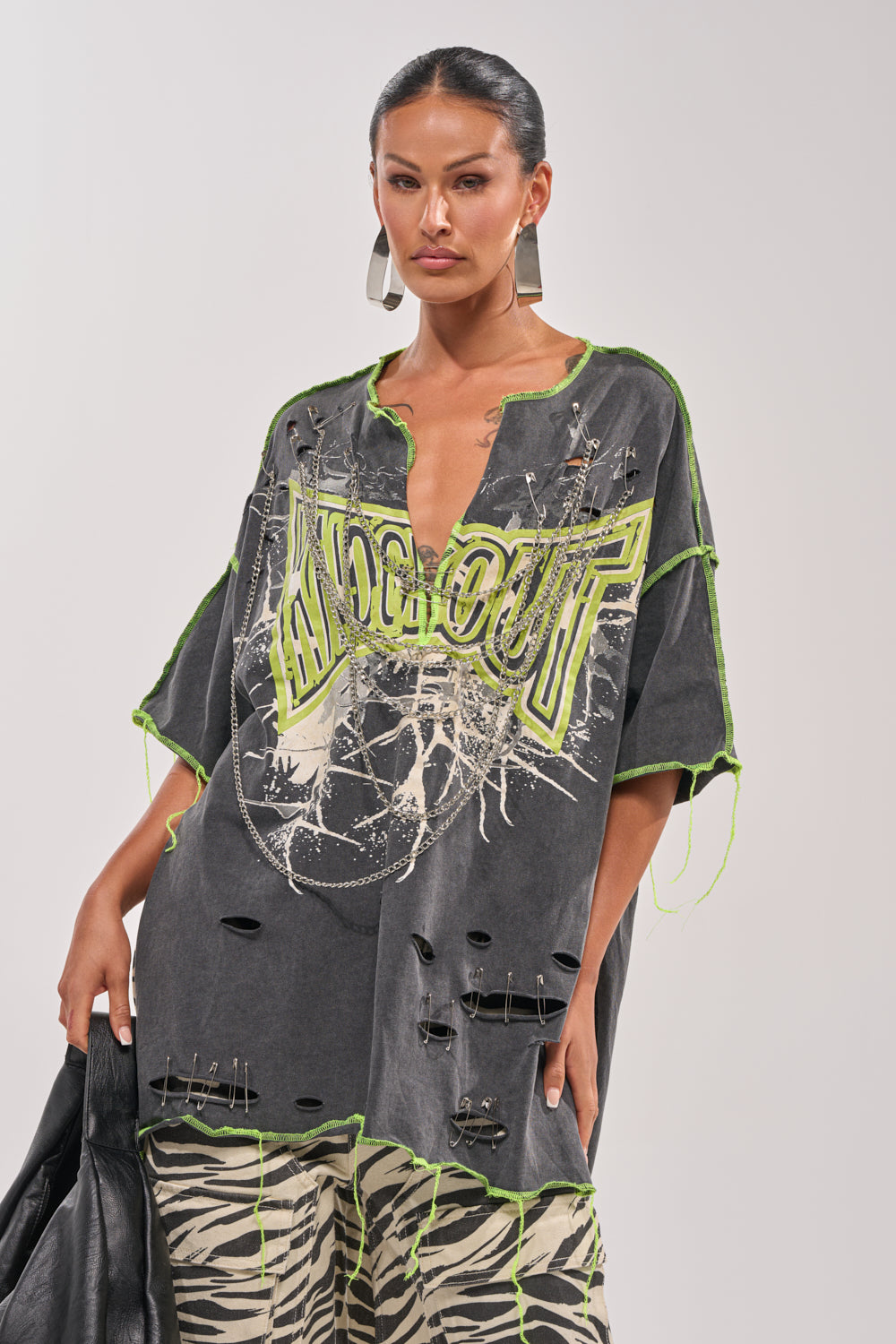 A woman models the KNOCKOUT OVERSIZED TEE, featuring distressed details, neon green stitching, safety pins, and bold “KNOCKOUT” text. She completes her streetwear look with zebra-print pants, hoop earrings, and a black bag. 0