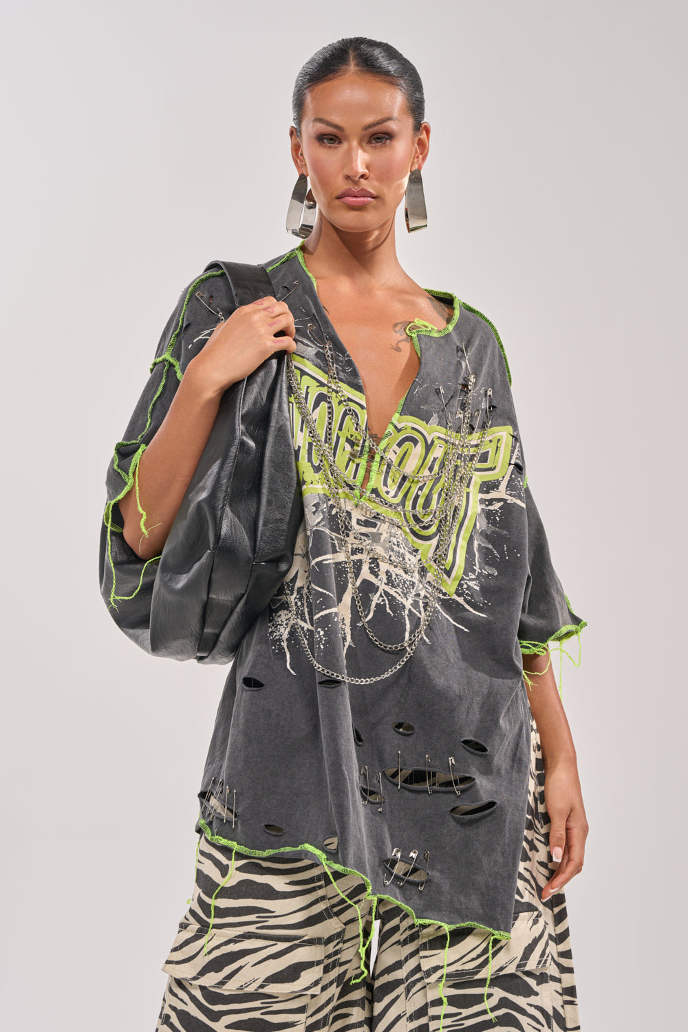 A woman poses confidently in streetwear, wearing the KNOCKOUT OVERSIZED TEE with neon green accents, zebra print pants, statement earrings, and a black bag with chain detail against a plain light background. 1