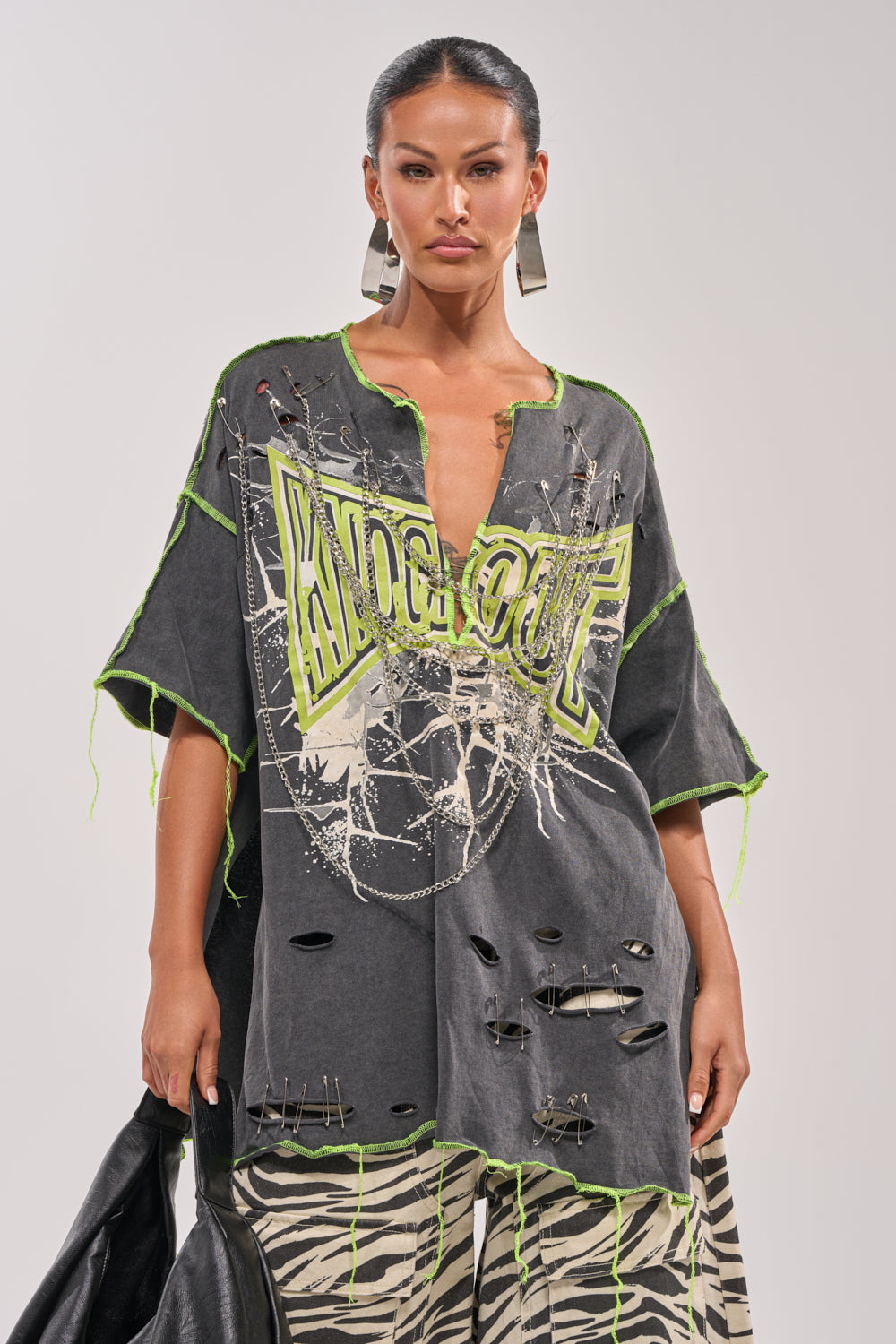 Wearing the KNOCKOUT OVERSIZED TEE—an oversized distressed graphic top with neon green trim and bold "KNOCKOUT" text—a woman pairs it with zebra print pants, geometric earrings, and holds a black jacket for a striking streetwear look. 2