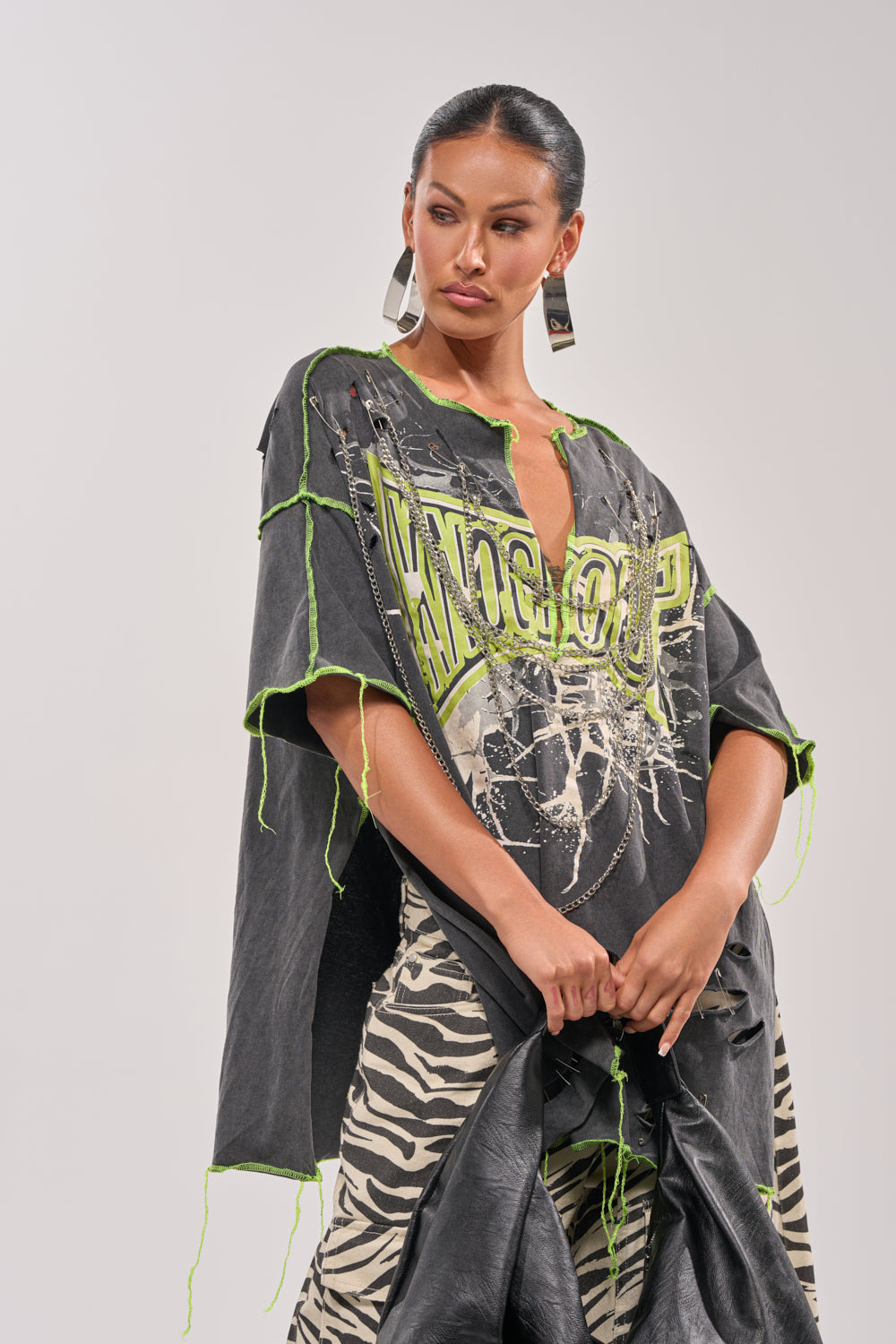 A woman with slicked-back hair and bold earrings rocks a streetwear look in the KNOCKOUT OVERSIZED TEE—an oversized black top with neon green stitching and distressed graphics—paired with zebra-print pants against a plain light background. 3