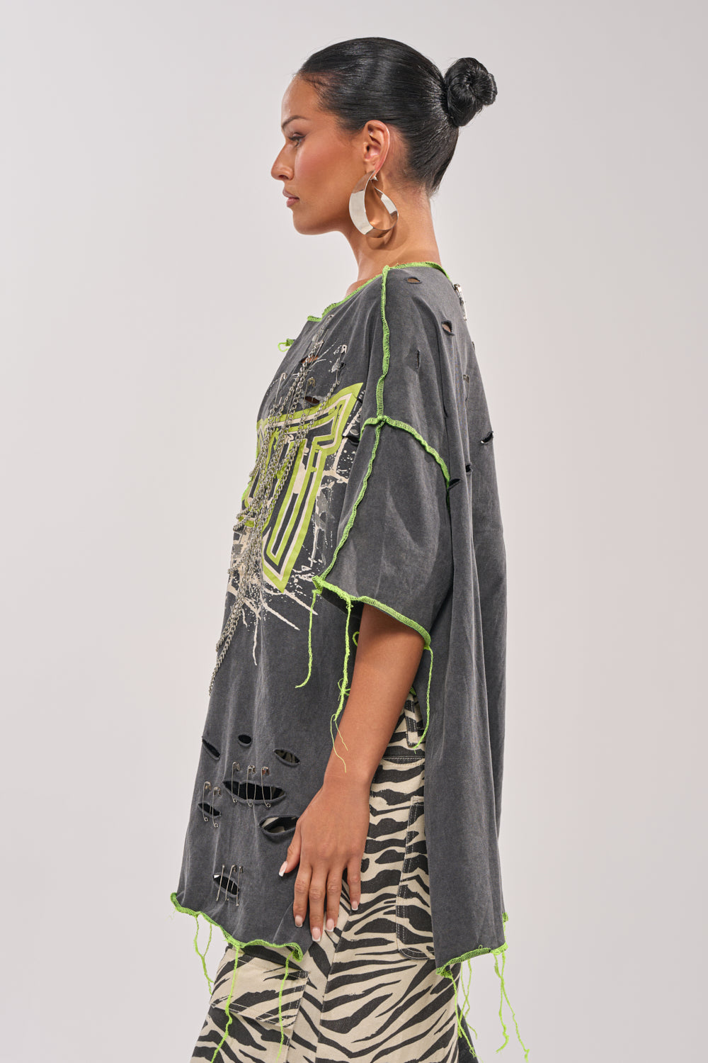 A woman in profile wears the KNOCKOUT OVERSIZED TEE with neon green accents, large hoop earrings, and a black-and-white zebra-print skirt. Her hair is styled in a neat bun, showcasing an edgy streetwear vibe against a plain background. 4