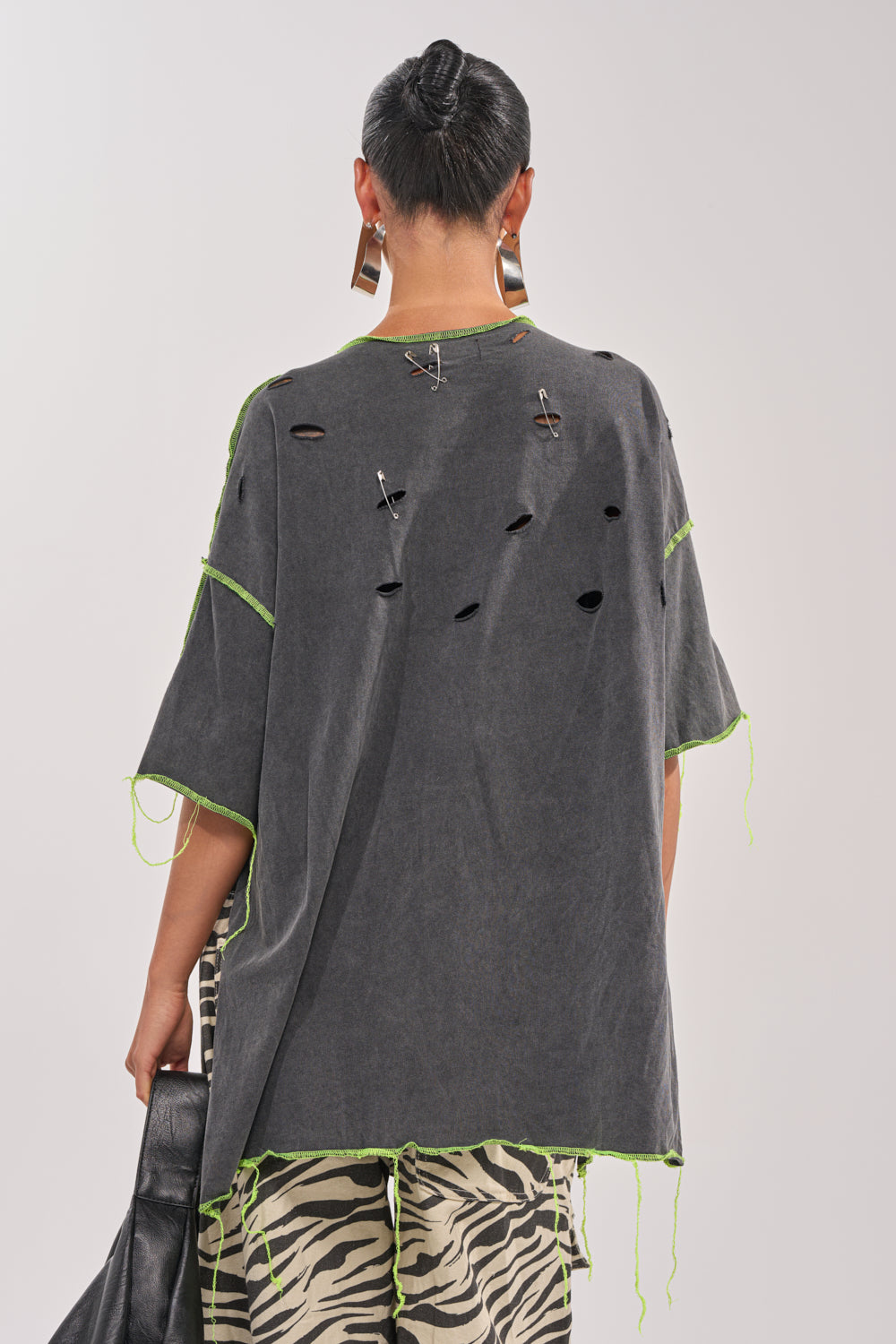 A person with short hair in a bun wears the KNOCKOUT OVERSIZED TEE featuring neon green stitching, holes, chains, and safety pins on the back, paired with zebra-print pants and geometric earrings for a bold streetwear look. 5