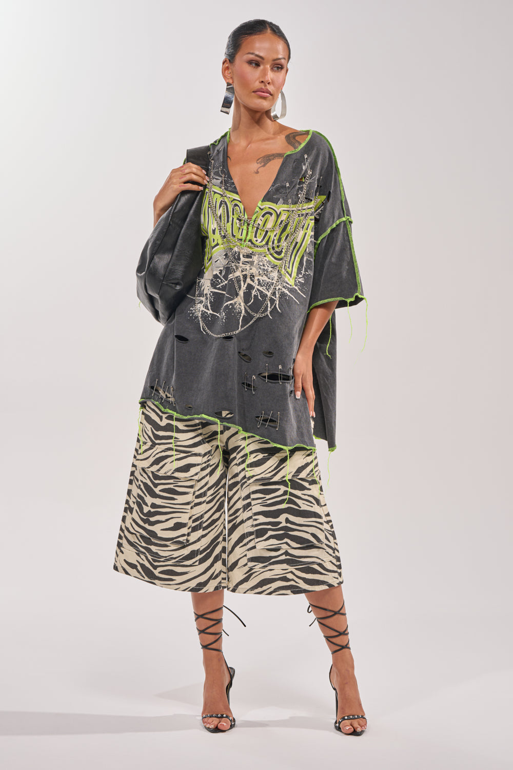 A woman stands confidently in streetwear, wearing the KNOCKOUT OVERSIZED TEE with neon green accents, zebra-print wide-leg pants, lace-up heels, hoop earrings, and a black chain-detailed handbag that highlights her visible tattoos. 6