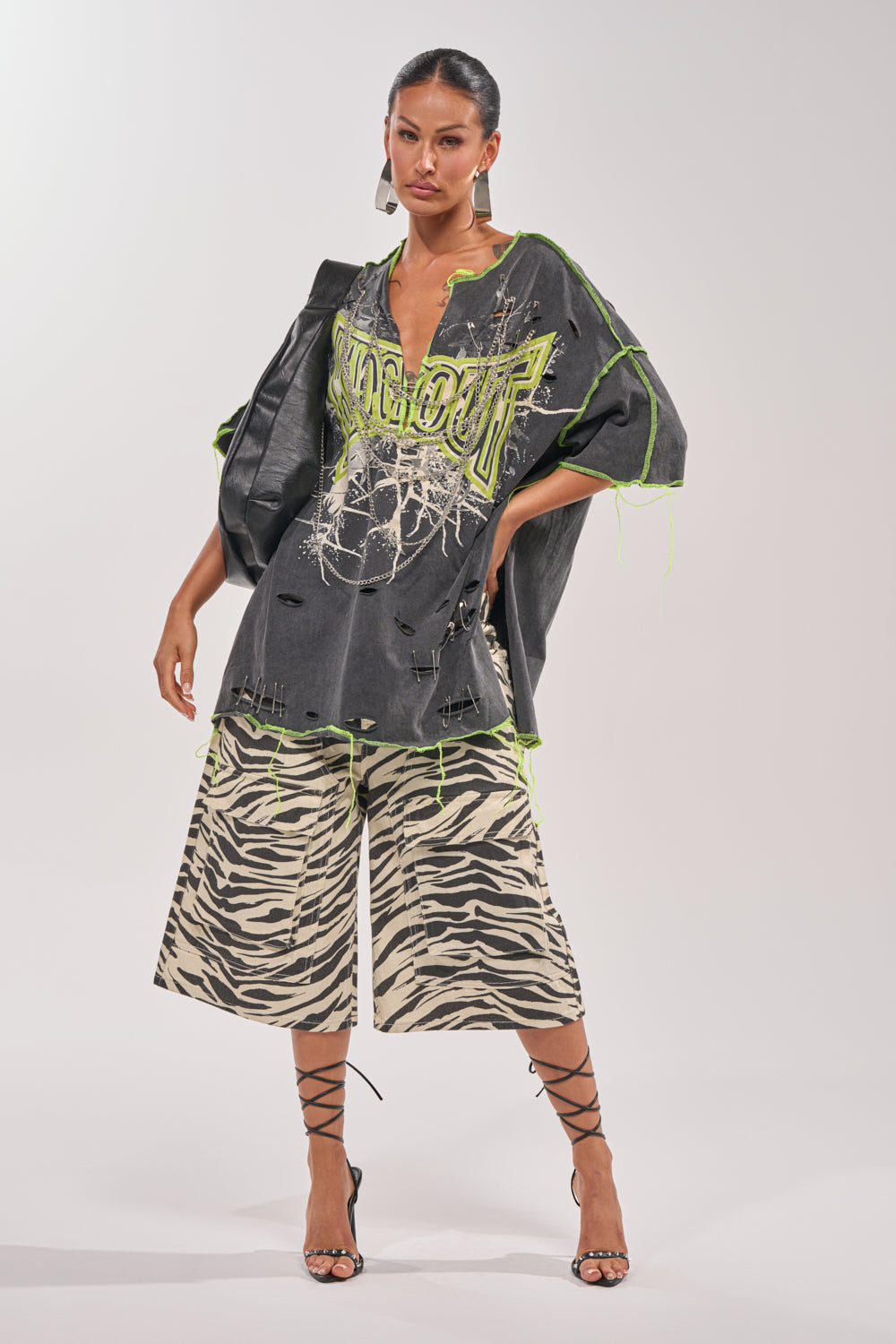 A woman rocks the KNOCKOUT OVERSIZED TEE with neon green and chain details, pairing it with wide zebra-print shorts. Strappy black heels, a large bag, and slicked-back hair finish her standout streetwear vibe. 7