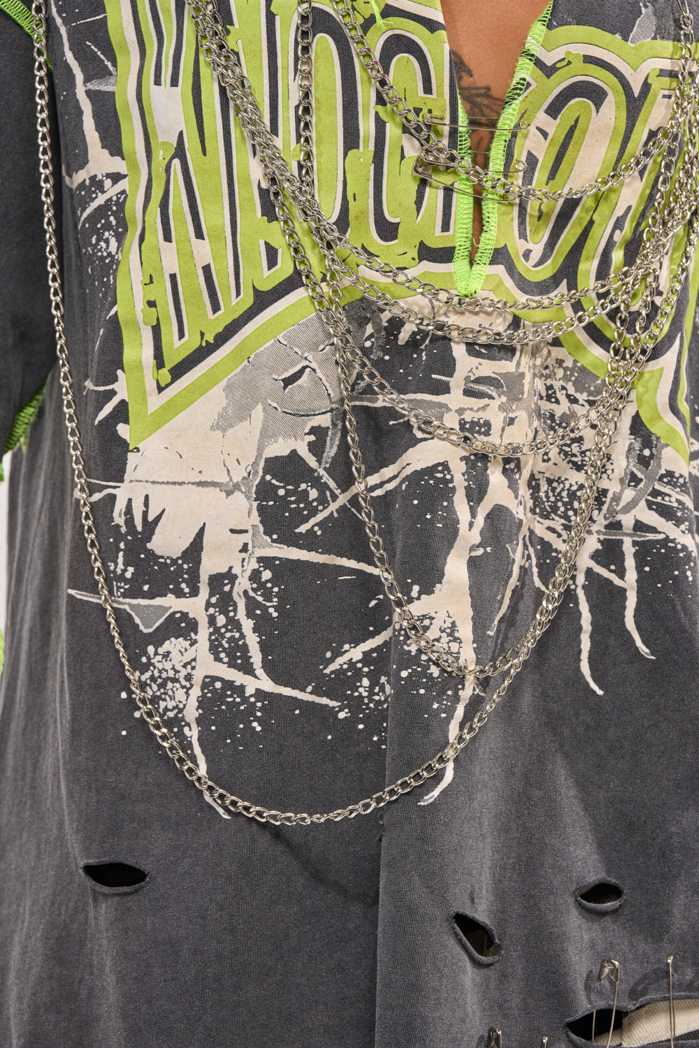 A close-up of the KNOCKOUT OVERSIZED TEE in grey, showcasing neon green and white streetwear graphics, splatter details, distressed holes, and silver chain accents layered across the chest. 8