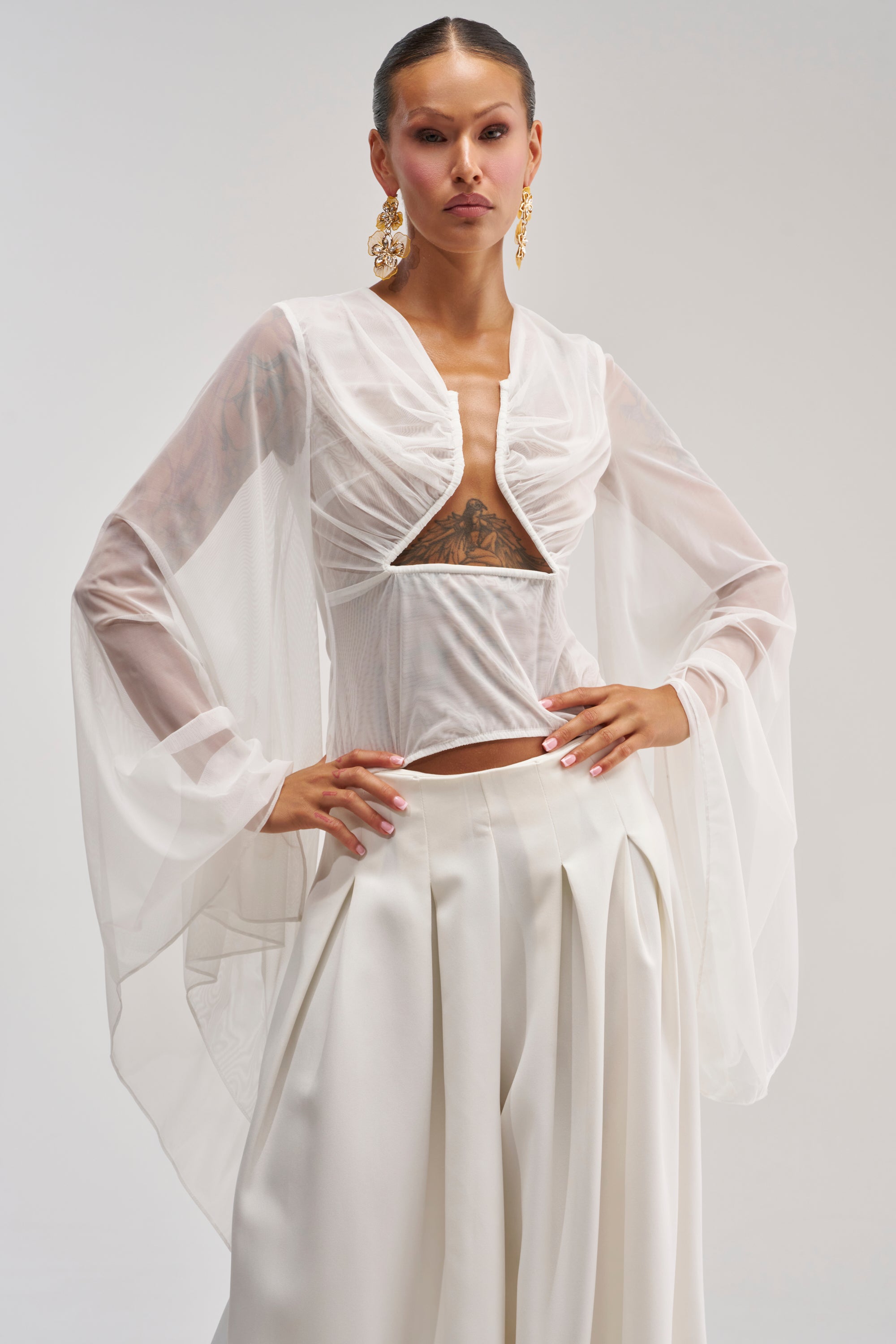 A person wearing the CELESTE CUTOUT DRAPE TOP IN WHITE, which features sheer chiffon sleeves and a structured bust, pairs it with high-waisted, pleated white pants while posing confidently against a light gray background.