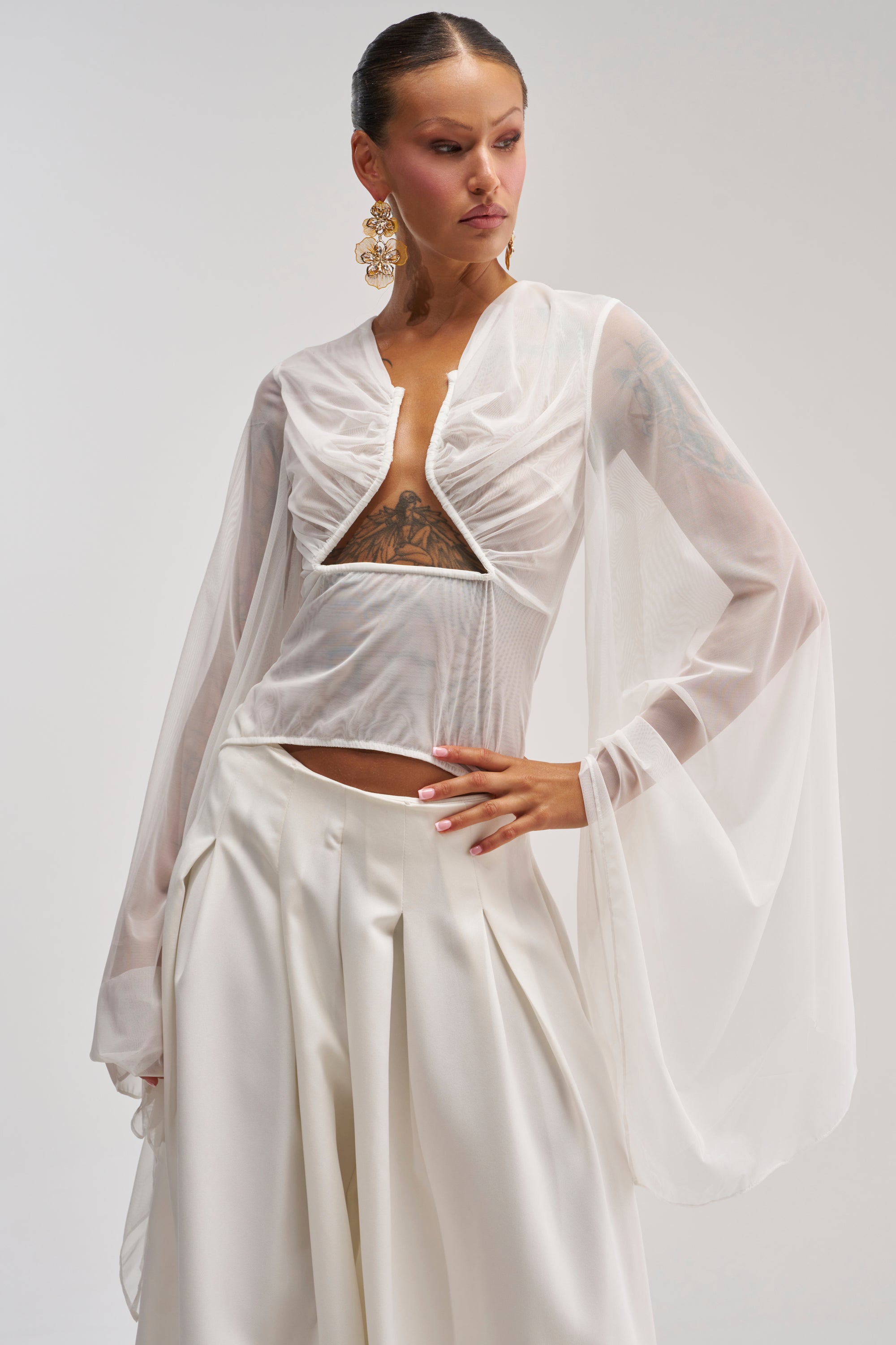 Wearing the CELESTE CUTOUT DRAPE TOP IN WHITE with sheer chiffon sleeves, a woman stands confidently in wide-leg white pants. She accessorizes with large, ornate earrings, and her chest and arm tattoos are visible.