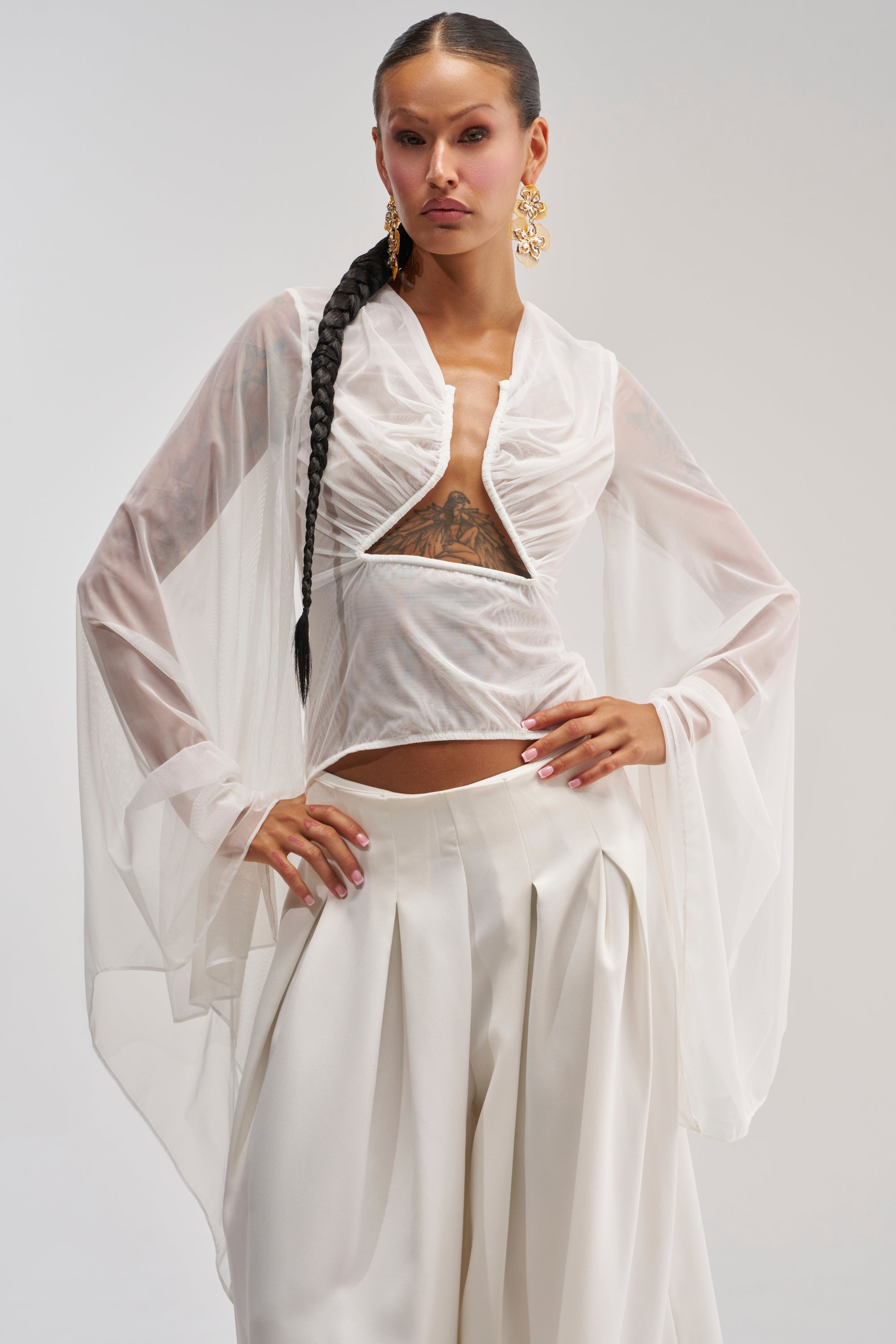A woman poses in the CELESTE CUTOUT DRAPE TOP IN WHITE, featuring sheer chiffon sleeves and paired with wide-legged white pants. She wears her hair in a long braid, accessorizes with gold earrings, and has a visible tattoo beneath her top, standing against a plain background.