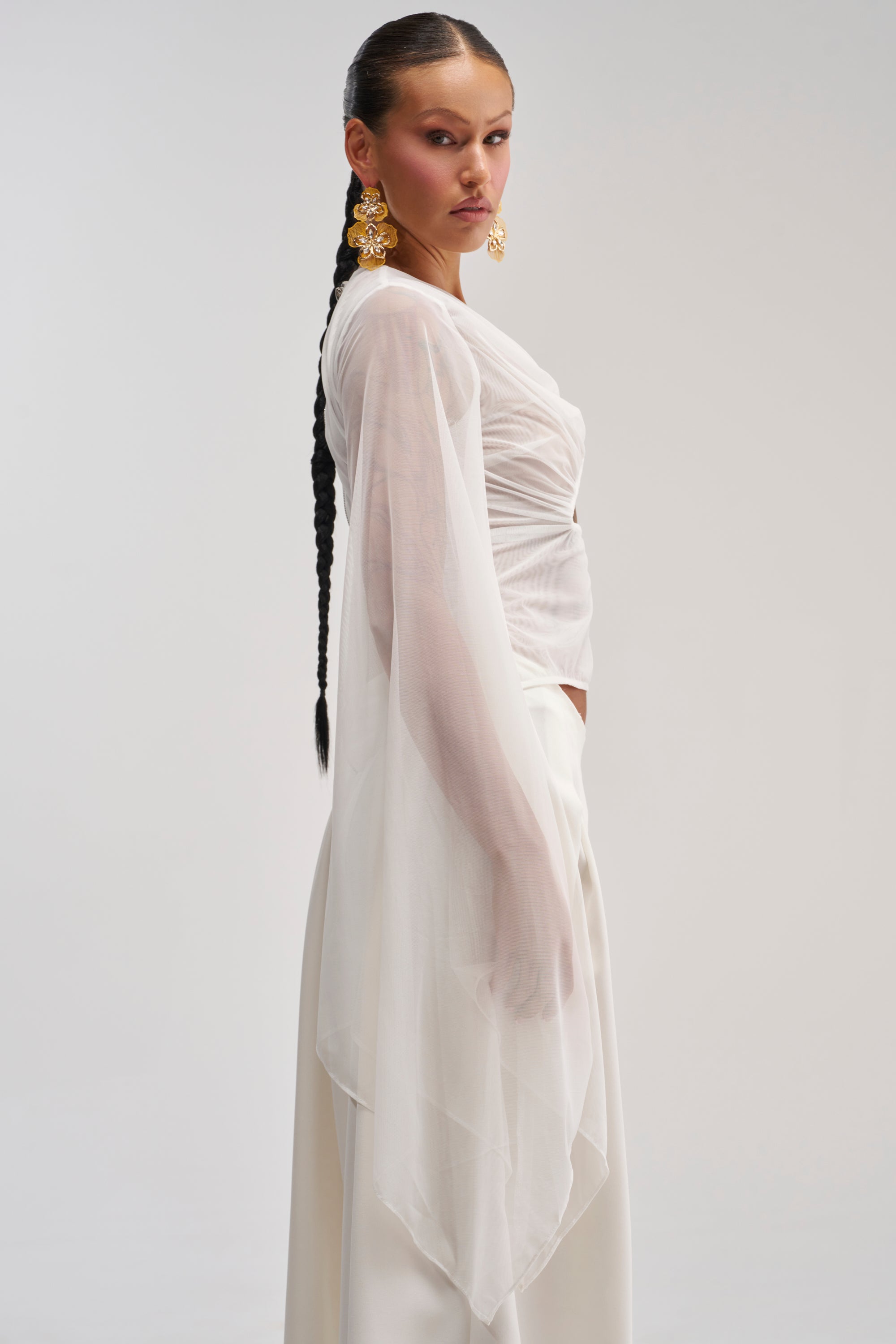 Woman with a long braid, wearing statement gold earrings and the CELESTE CUTOUT DRAPE TOP IN WHITE featuring sheer chiffon sleeves, stands in profile against a plain light background.