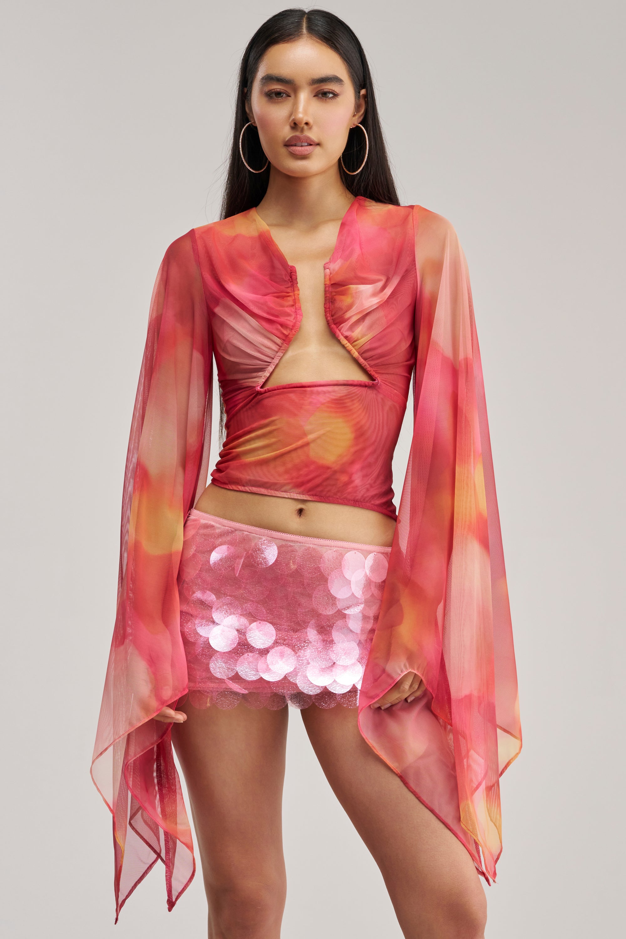 A woman stands facing forward against a plain light gray background, wearing the CELESTE CUTOUT DRAPE TOP—a sheer mesh top in flowy pink and orange tones with dramatic long sleeves—paired with a short pink sequined skirt.