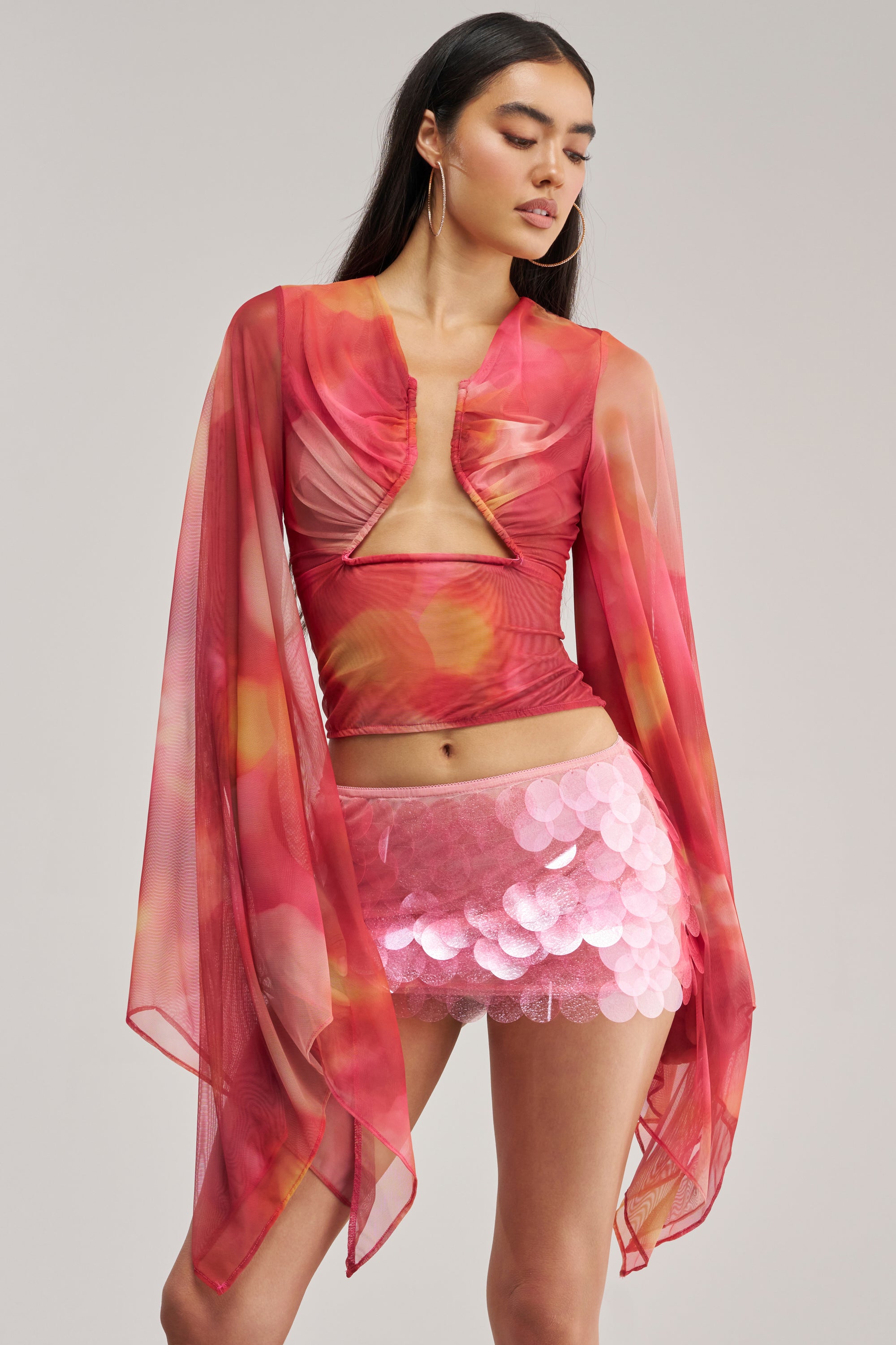 Wearing the CELESTE CUTOUT DRAPE TOP—a sheer mesh design in flowing pink and orange with dramatic sleeves and a cutout front—she pairs it with a short, shiny pink sequin skirt, gazing to the side against a plain background.